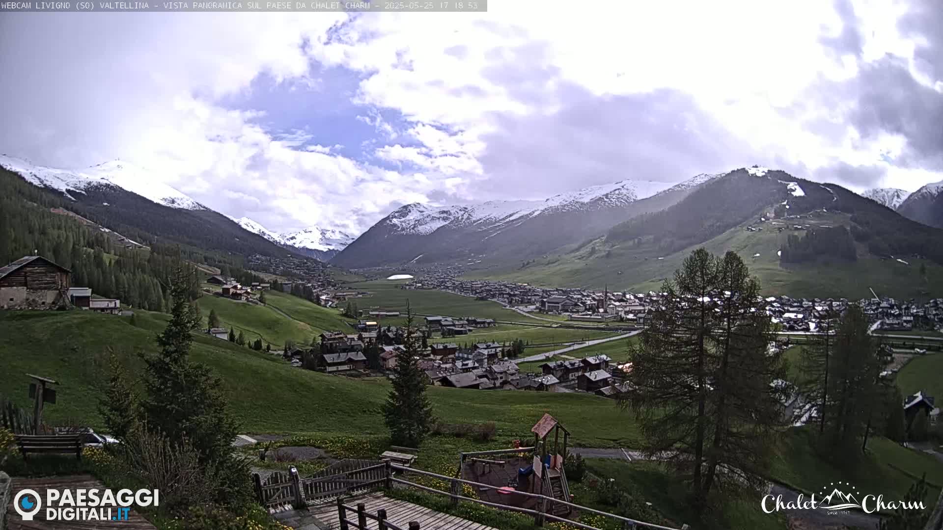 A partly cloudy day reveals a snow-capped mountain range overlooking a verdant valley town with a playground in the foreground.