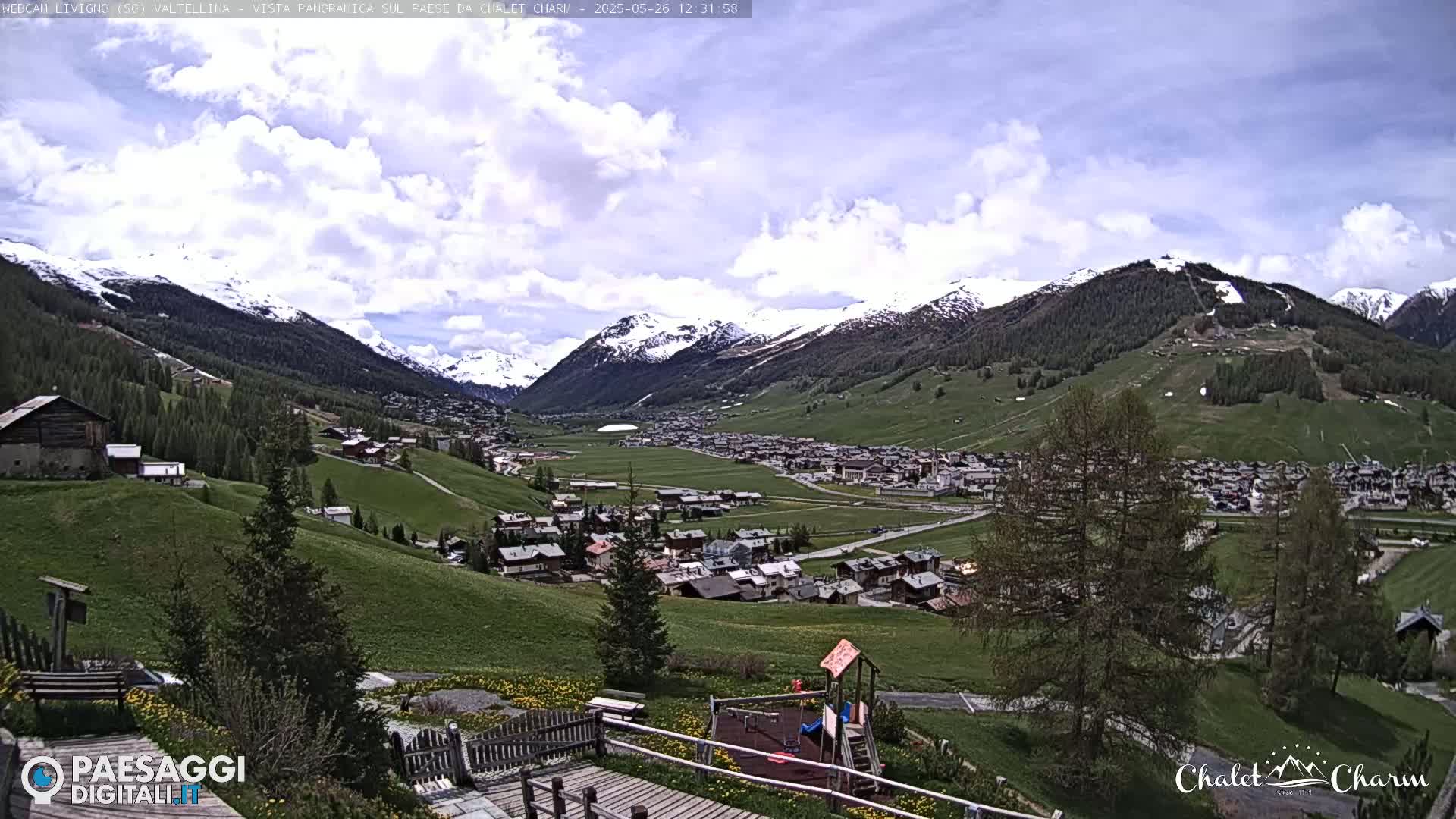 A partly cloudy day reveals a snow-capped mountain range overlooking a small village nestled in a green valley, with a playground visible in the foreground.