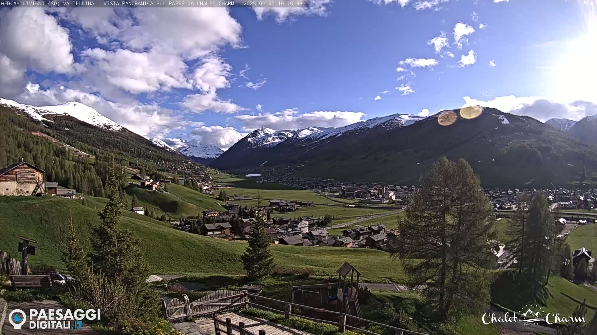 A partly sunny day reveals a mountainous valley town with snow-capped peaks in the background, green hills in the foreground, and a small playground visible.