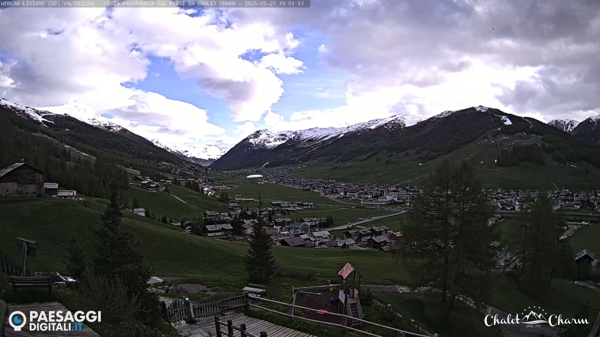 A partly cloudy afternoon reveals a snow-capped mountain range overlooking a valley town with green pastures and a playground.