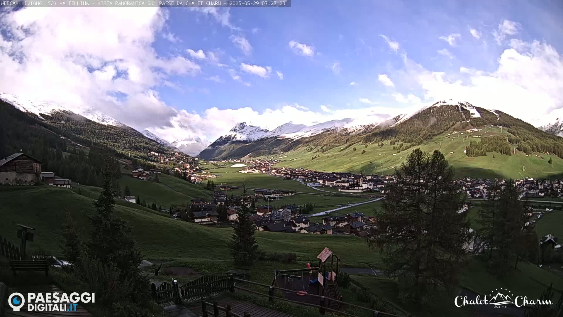 A partly cloudy, sunny day reveals a valley town nestled between snow-capped mountains and green hillsides.