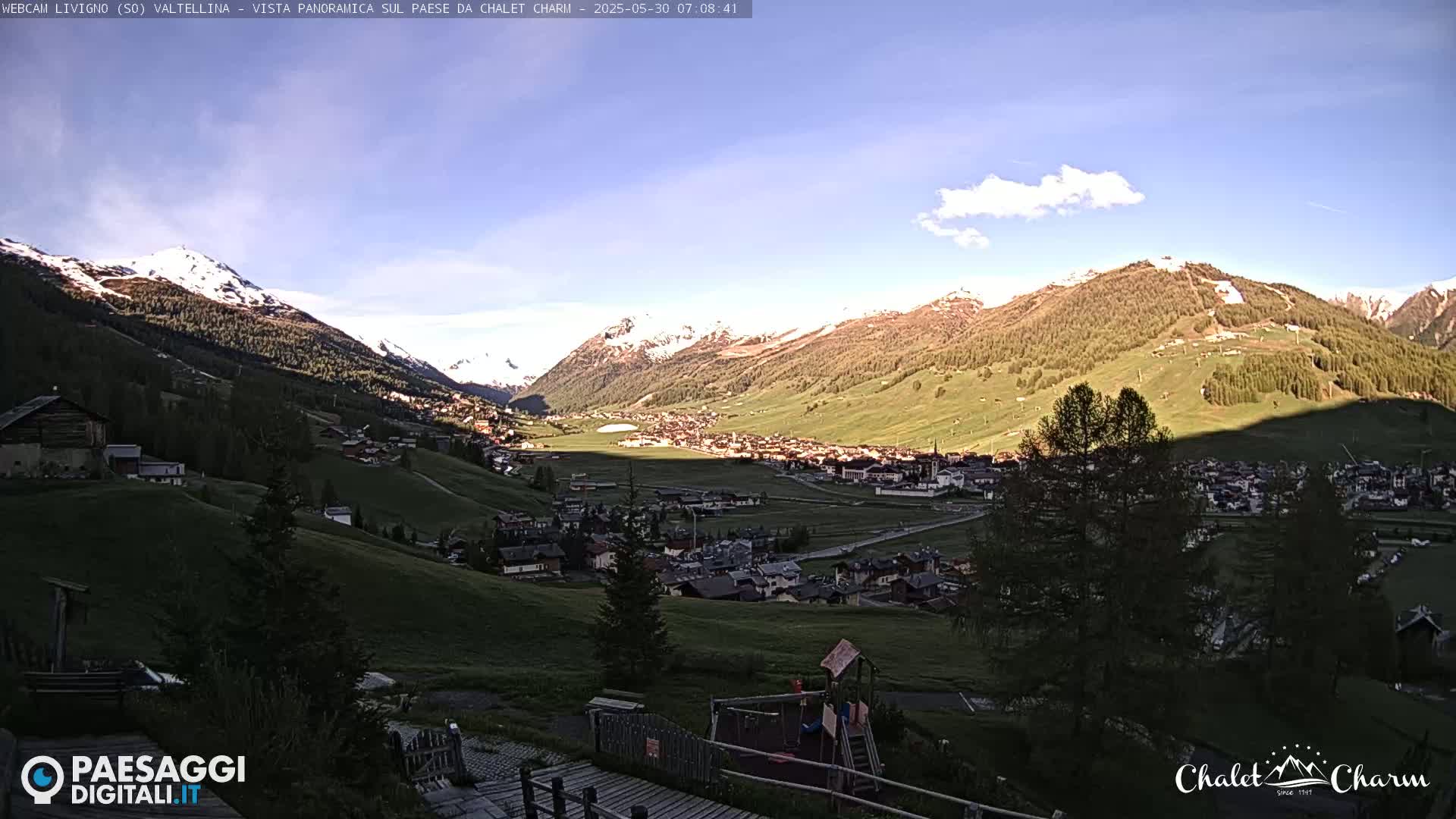 A picturesque mountain valley town is nestled between snow-capped mountains under a mostly clear, sunny sky.