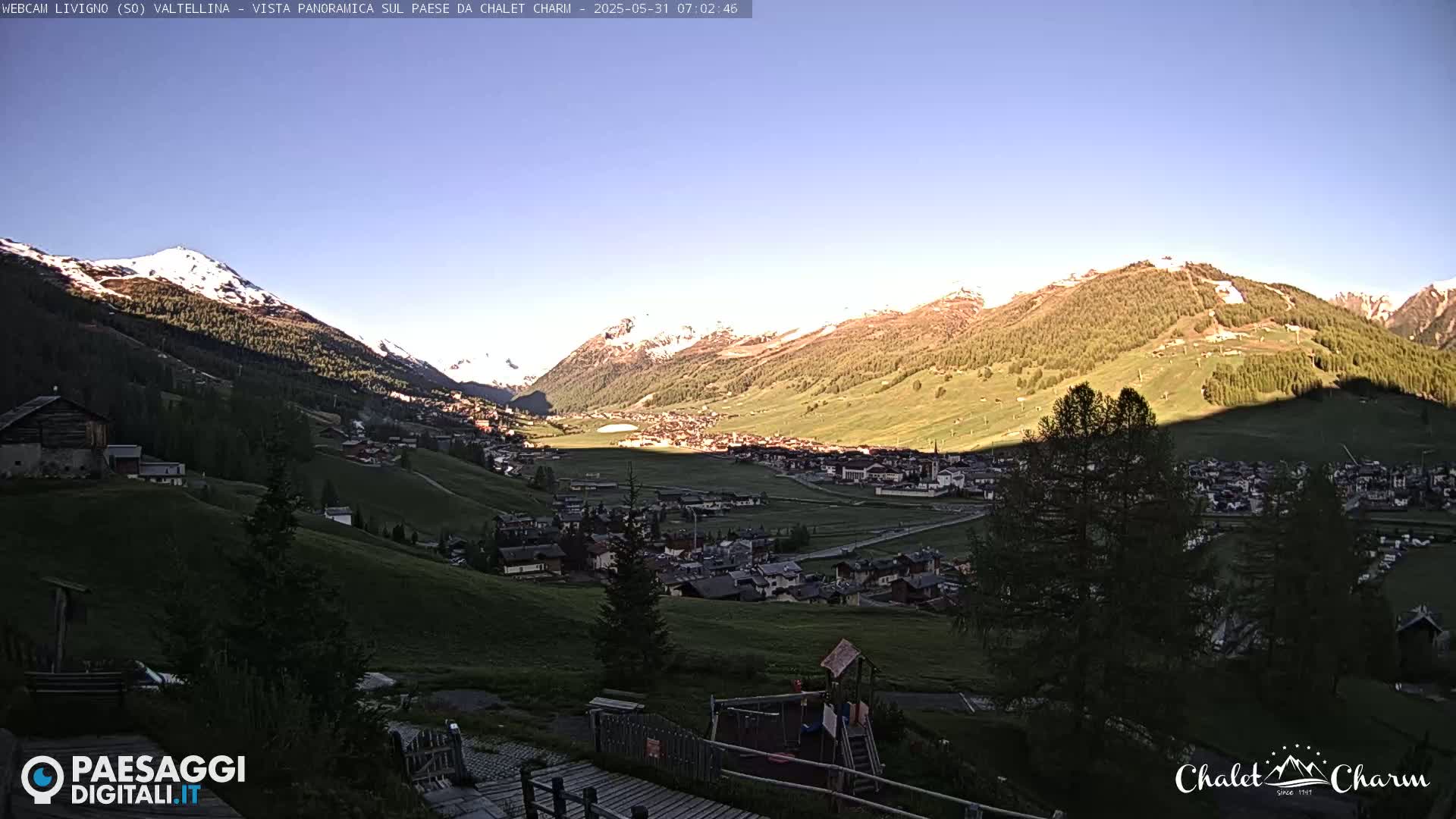A sunlit mountain valley town is nestled amongst green hills and snow-capped mountains under a clear blue sky.