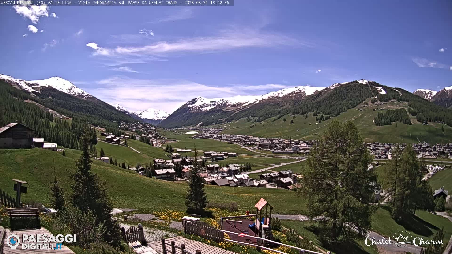 A sunny day reveals a picturesque village nestled in a valley surrounded by snow-capped mountains and lush green hills.