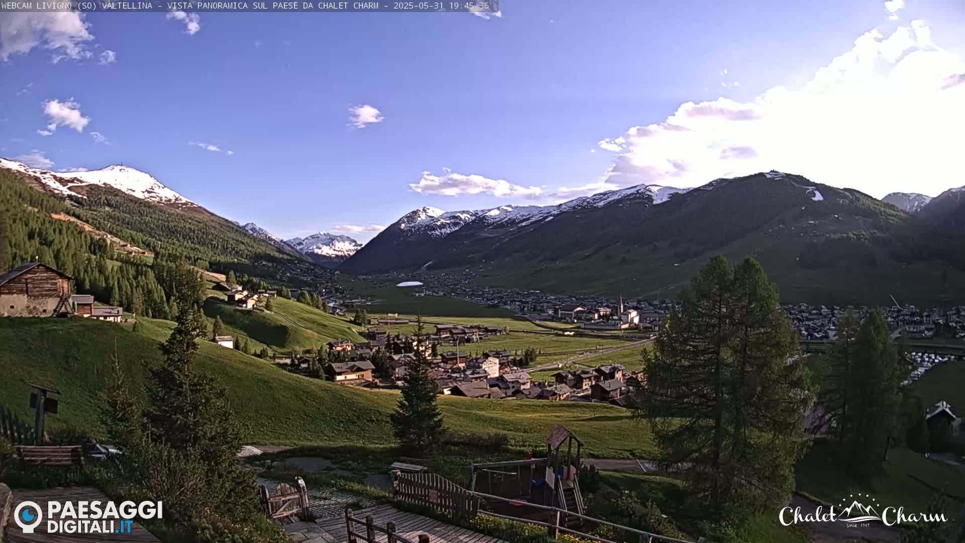 A picturesque valley town nestled among snow-capped mountains under a partly cloudy, bright sky.