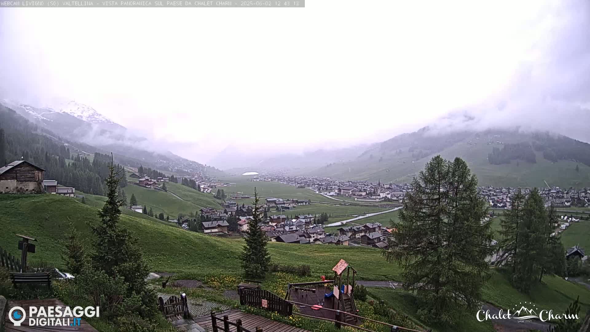 A misty, overcast view shows a valley town nestled among green hills and mountains, with a playground visible in the foreground.