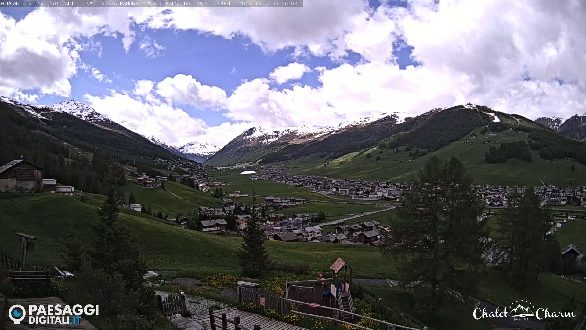 A partly cloudy day reveals a mountain valley town nestled among green hills and snow-capped mountains.
