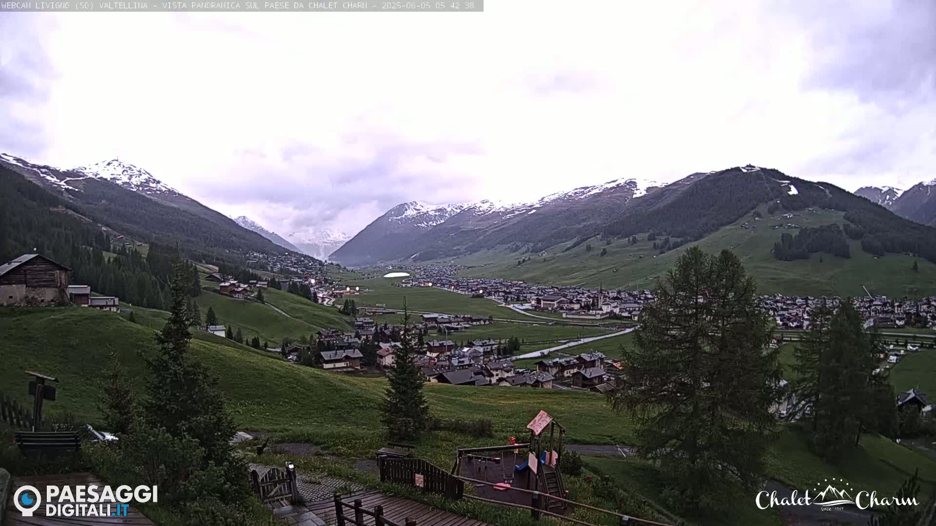 A long-range view of a village nestled in a valley between snow-capped mountains under an overcast sky.