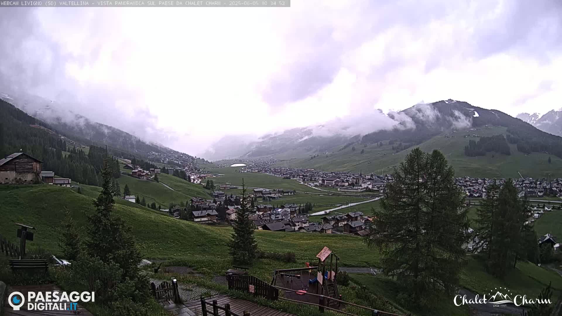 A mountain village nestled in a valley is partially obscured by low-hanging clouds under an overcast sky.