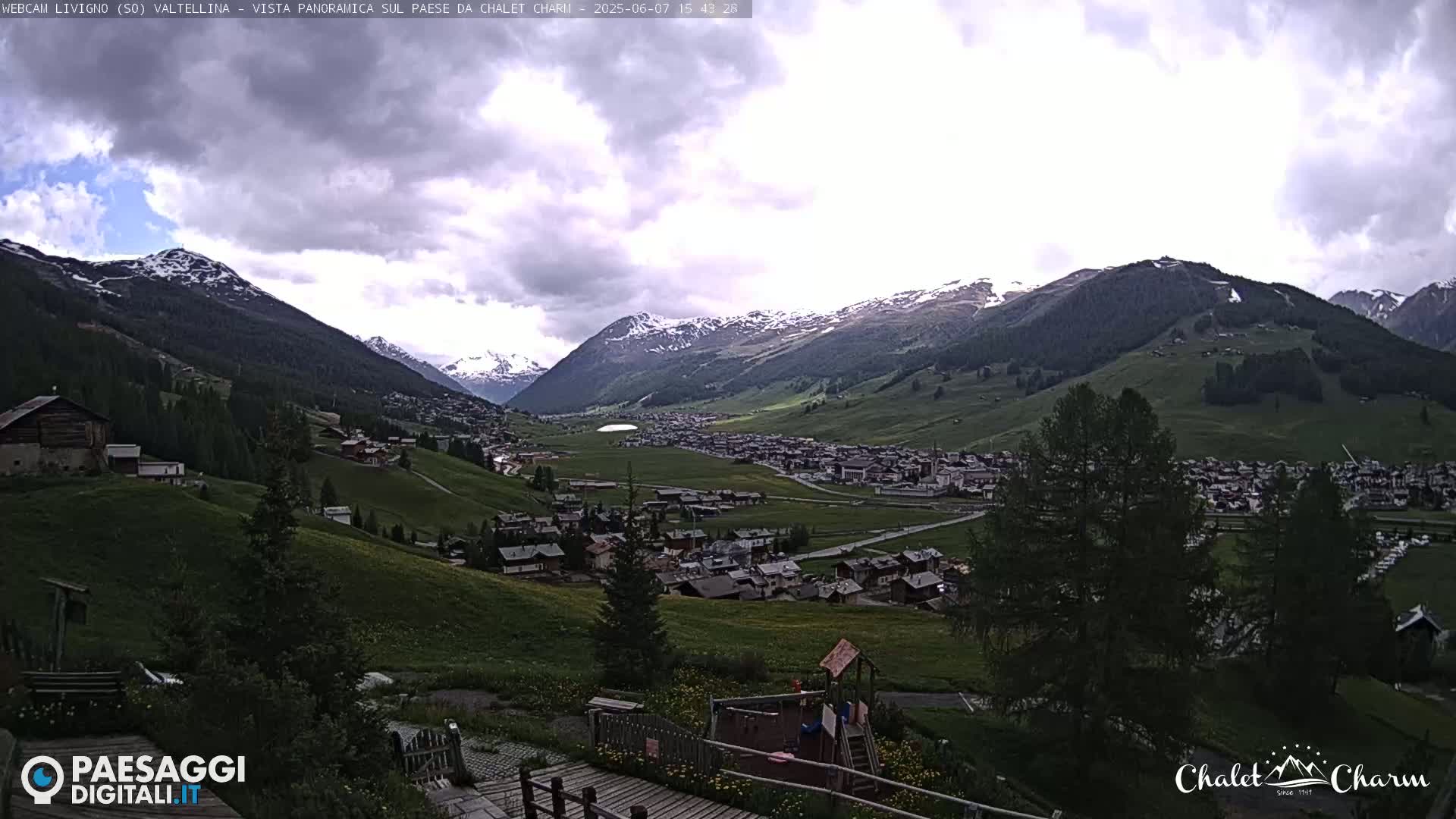 A panoramic view of a village nestled in a valley between snow-capped mountains under a partly cloudy sky.