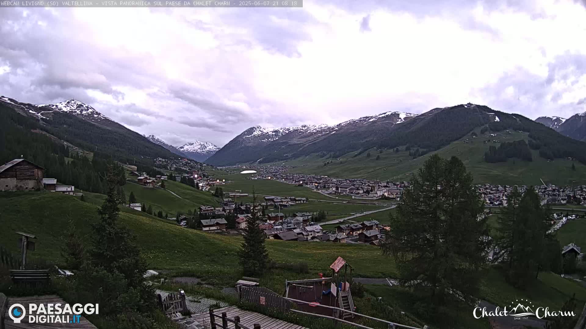 A mountain village nestled in a valley between snow-capped mountains under a partly cloudy sky.
