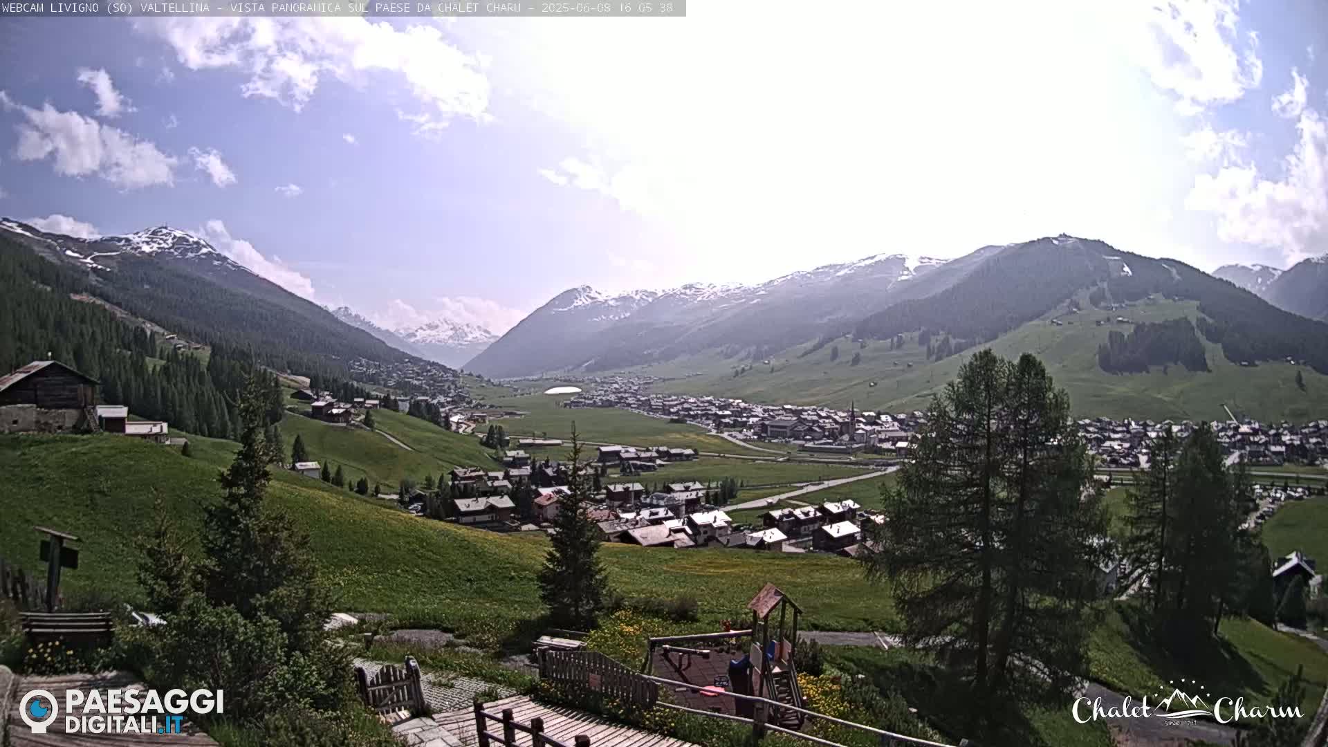 A partly sunny view of a village nestled in a valley surrounded by snow-capped mountains.