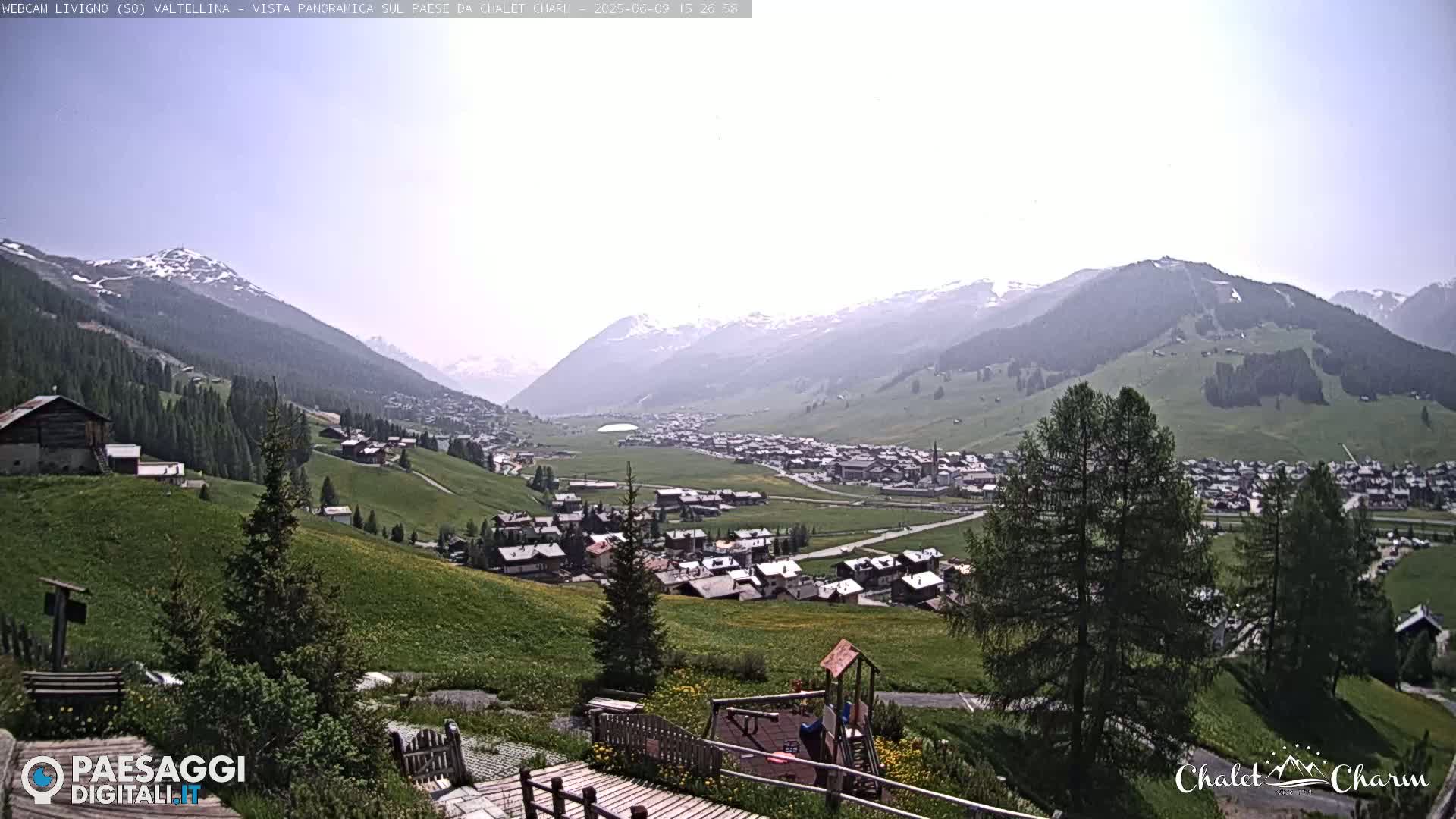 A sunny, panoramic view of a village nestled in a valley surrounded by snow-capped mountains and green hills.