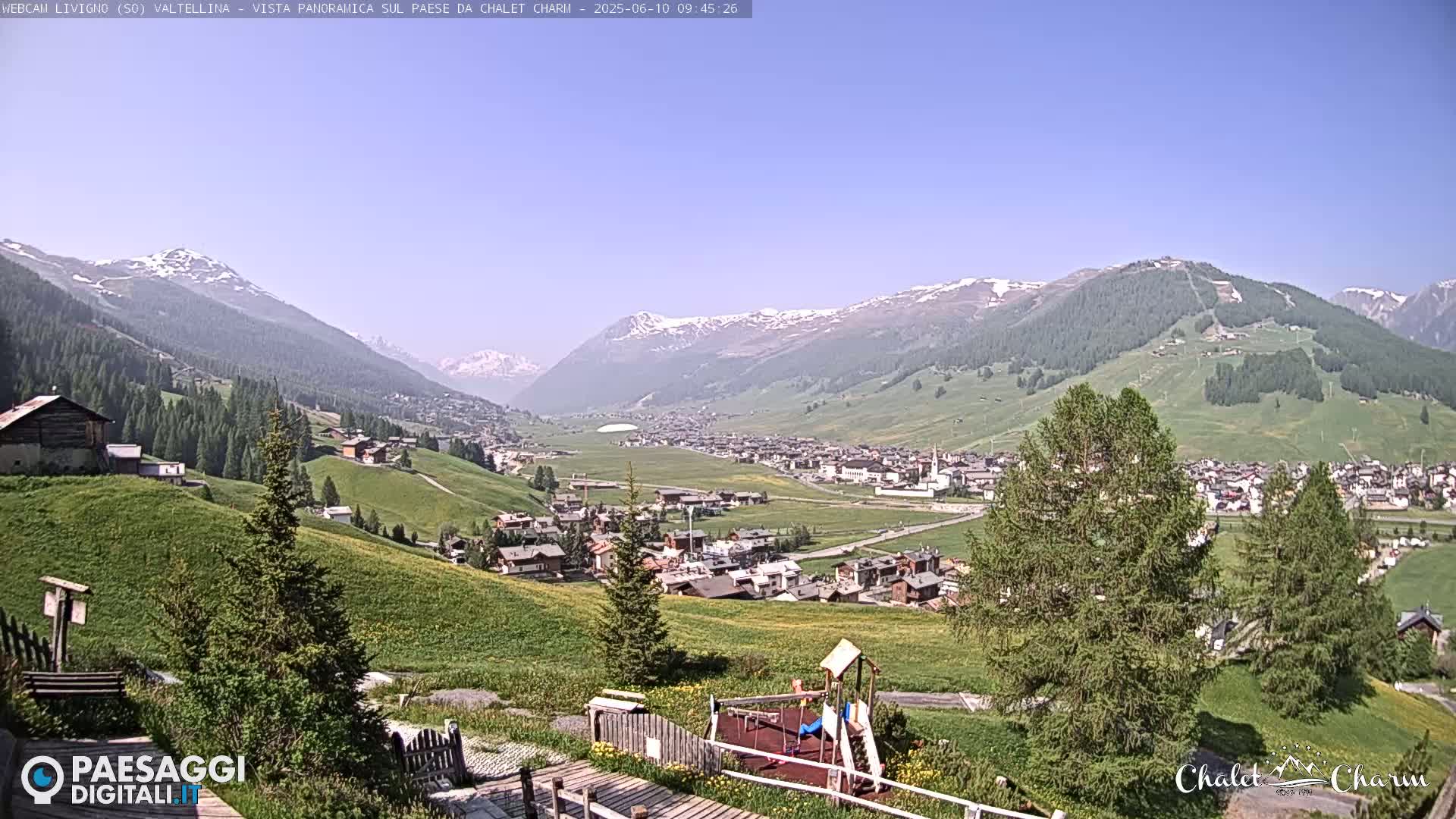 A sunny view of a valley town nestled among snow-capped mountains and green hills, with a playground visible in the foreground.