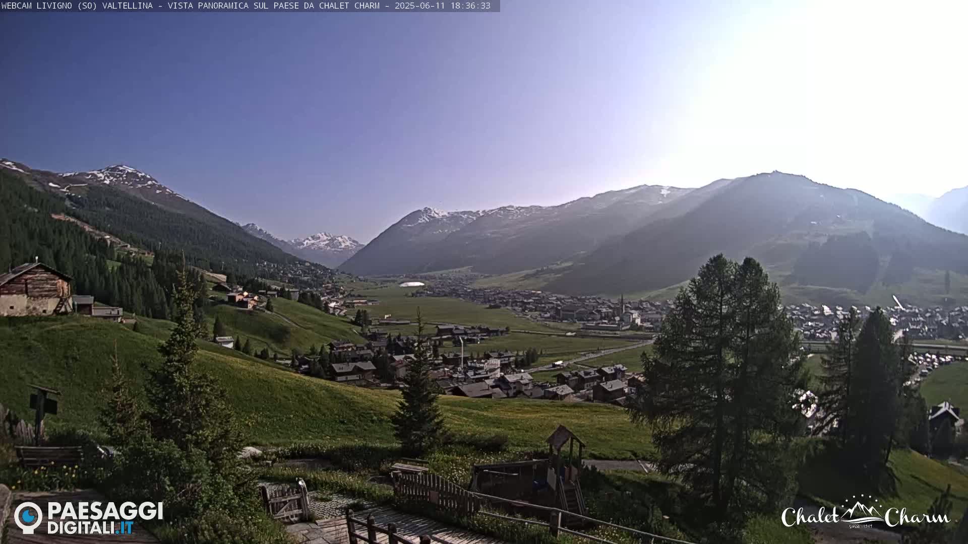 A sunny, panoramic view of a valley town nestled among green hills and snow-capped mountains.