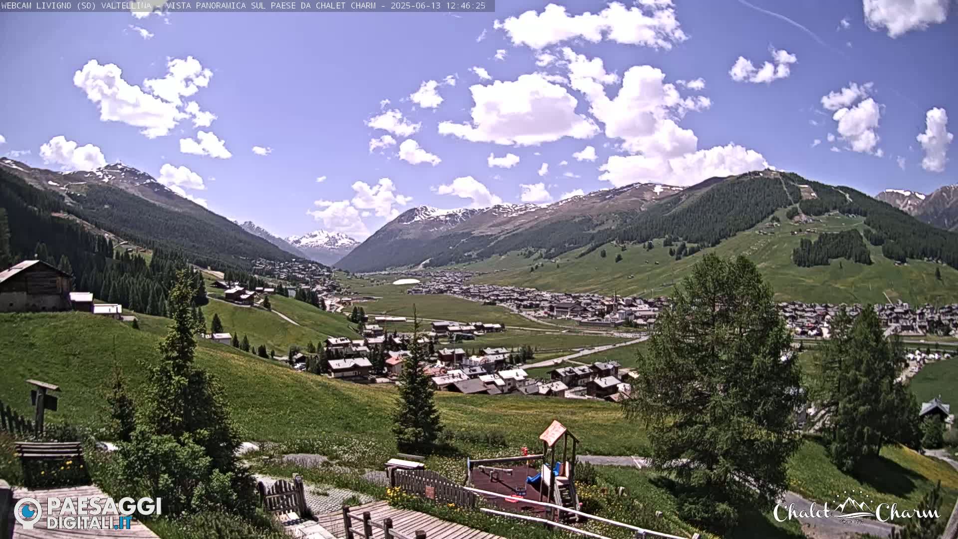 A partly cloudy day reveals a mountainous village nestled in a green valley, with a playground visible in the foreground.
