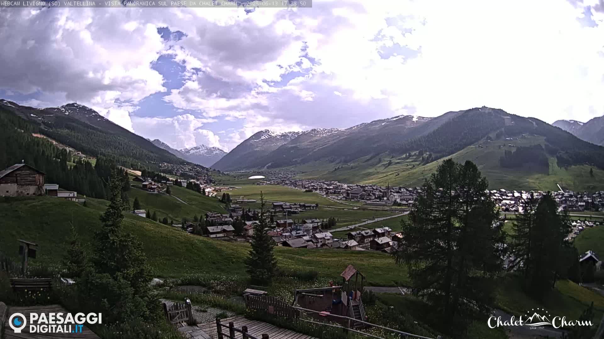 A partly cloudy day reveals a picturesque mountain valley village nestled among green hills and snow-capped mountains.