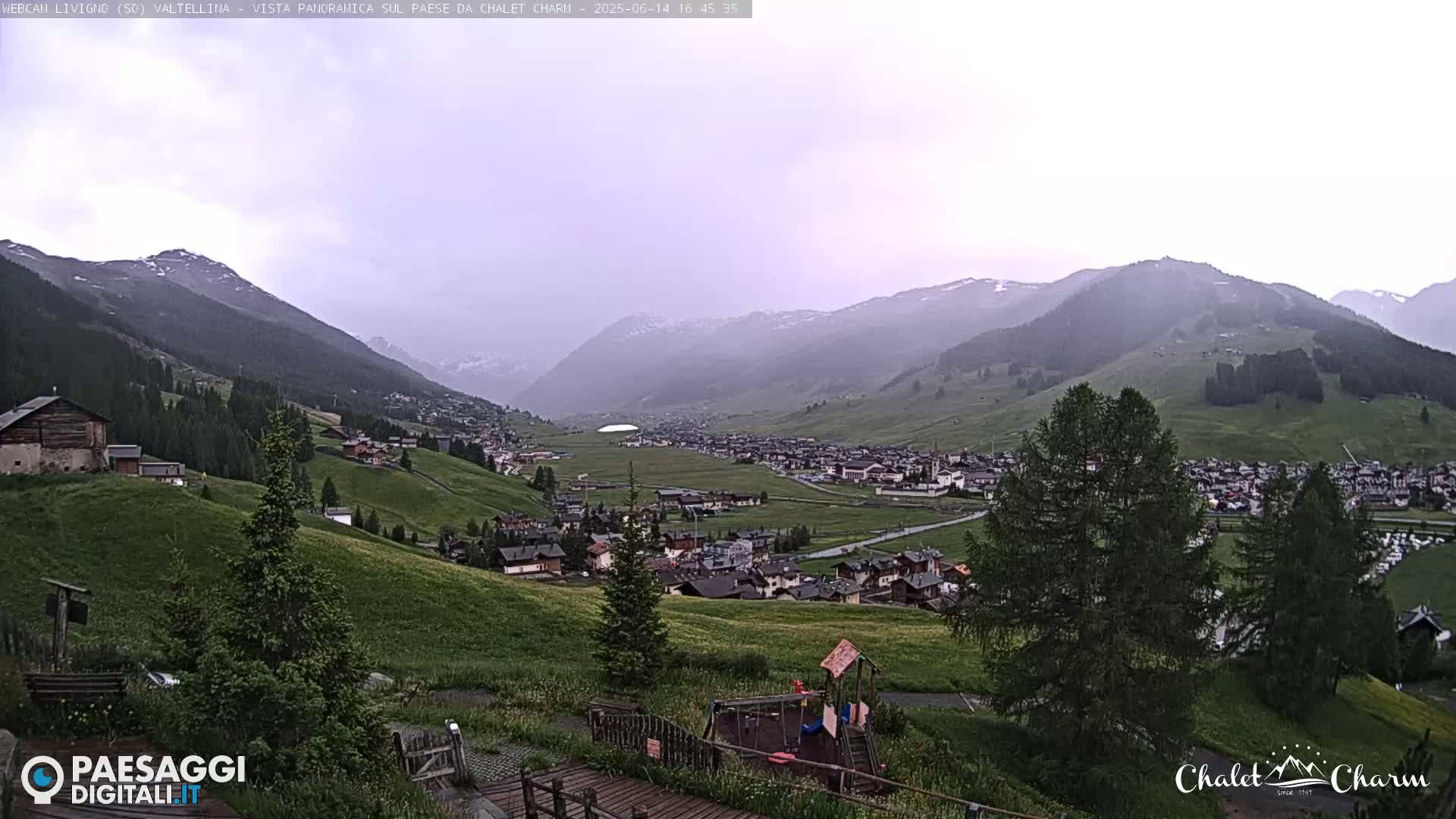A mountain village nestled in a valley under an overcast sky, with green hillsides and a playground visible in the foreground.