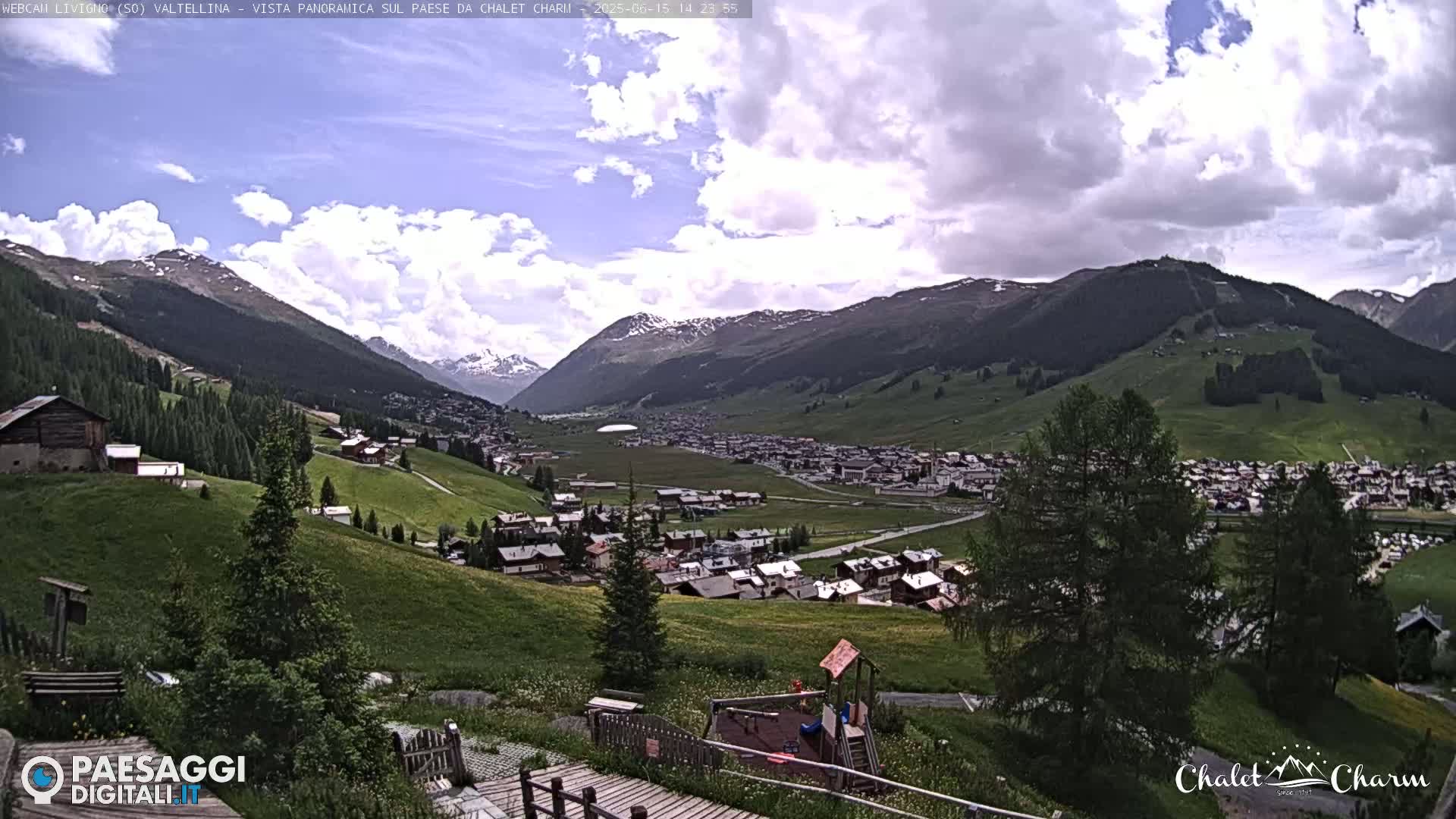 A panoramic view of a village nestled in a valley surrounded by mountains under a partly cloudy sky.