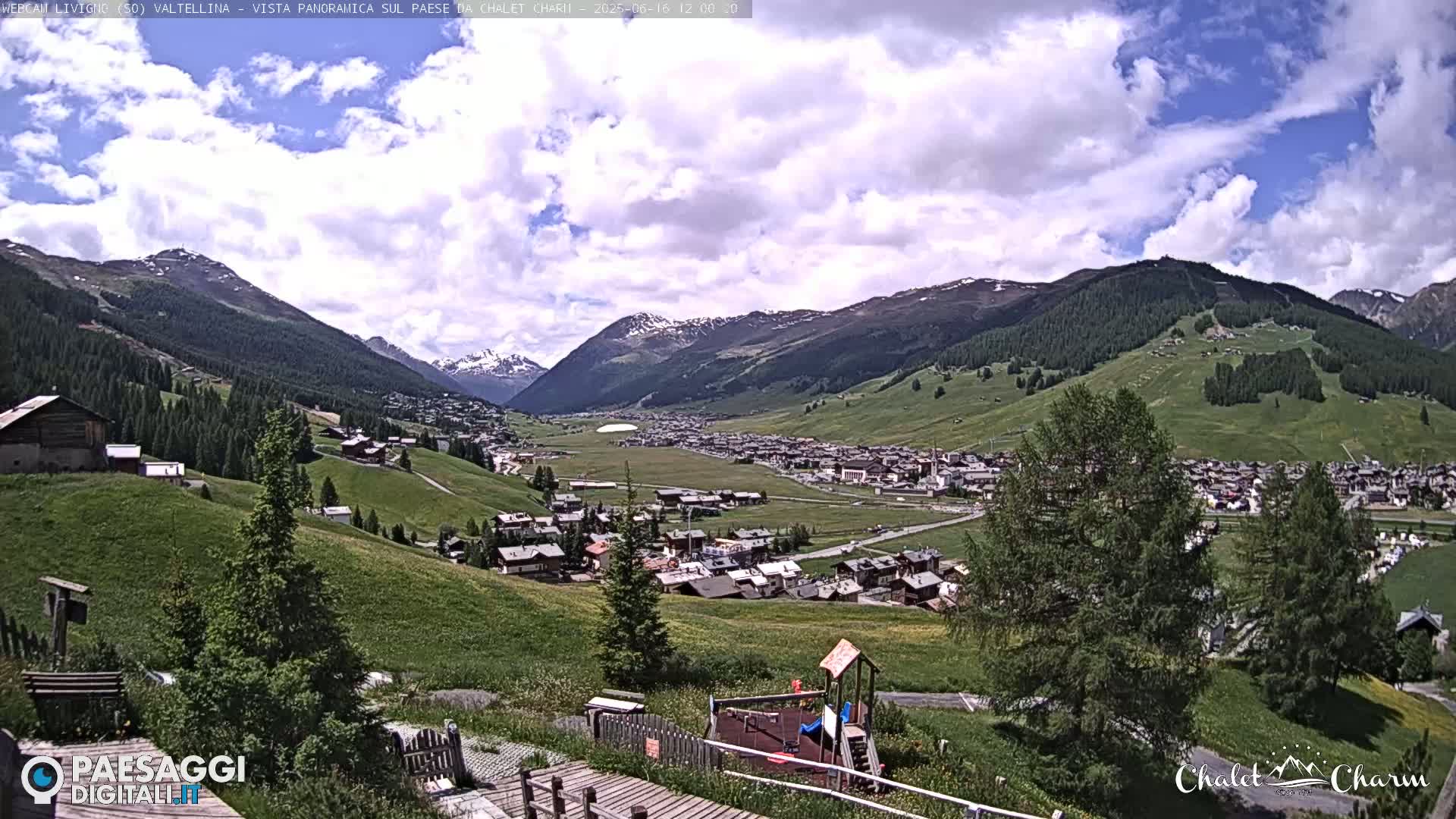 A partly cloudy day reveals a panoramic view of a valley town nestled among green hills and snow-capped mountains.