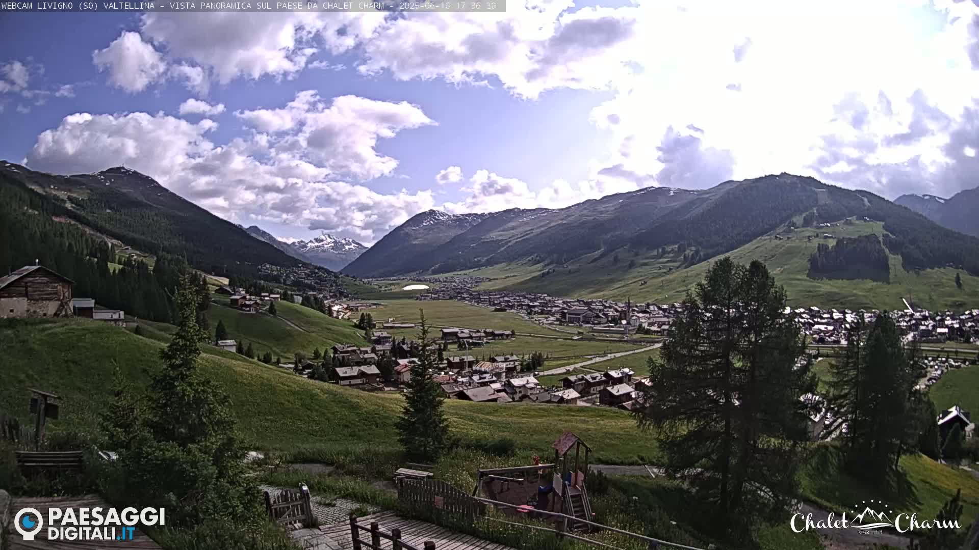 A partly cloudy day reveals a picturesque valley town nestled in a mountainous region, with green hillsides and a children's play area in the foreground.