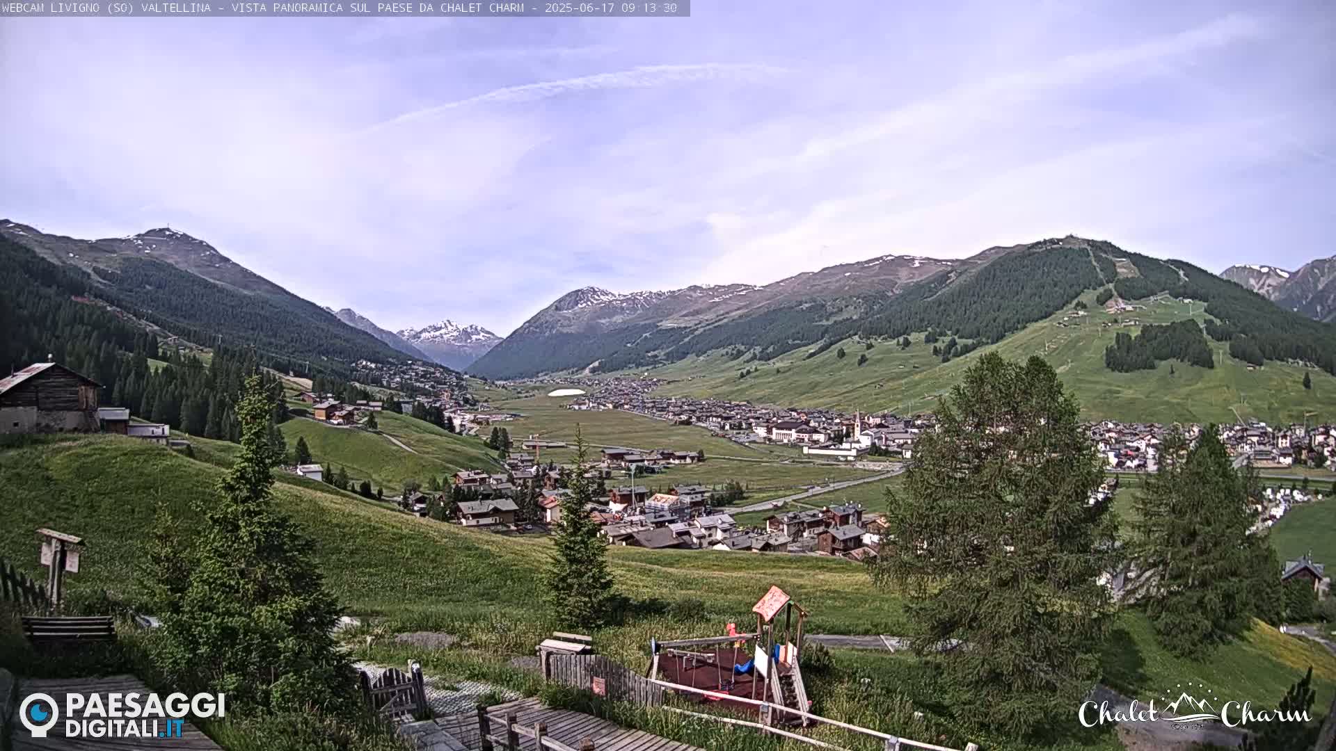 A panoramic view of a village nestled in a valley surrounded by green, rolling hills and mountains under a partly cloudy sky.