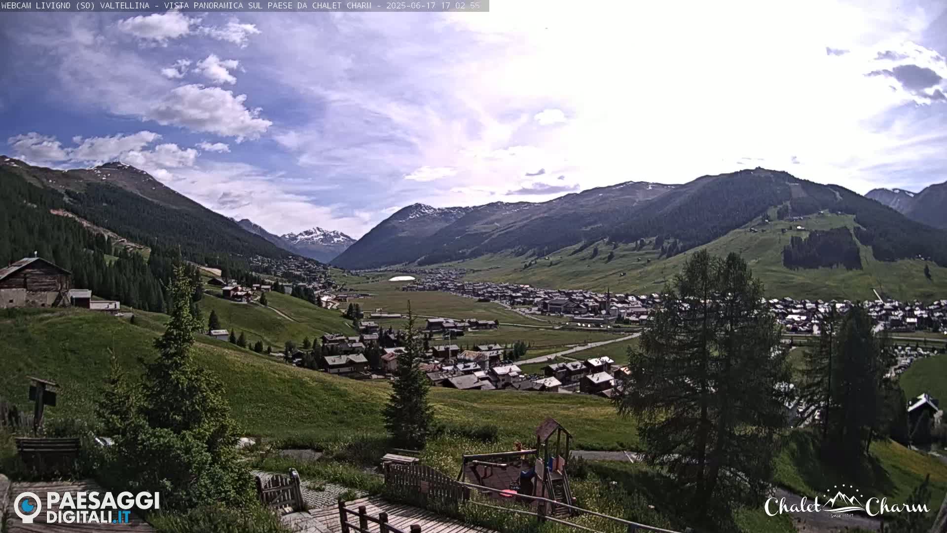 A partly cloudy day reveals a panoramic view of a village nestled in a valley surrounded by verdant hills and mountains.