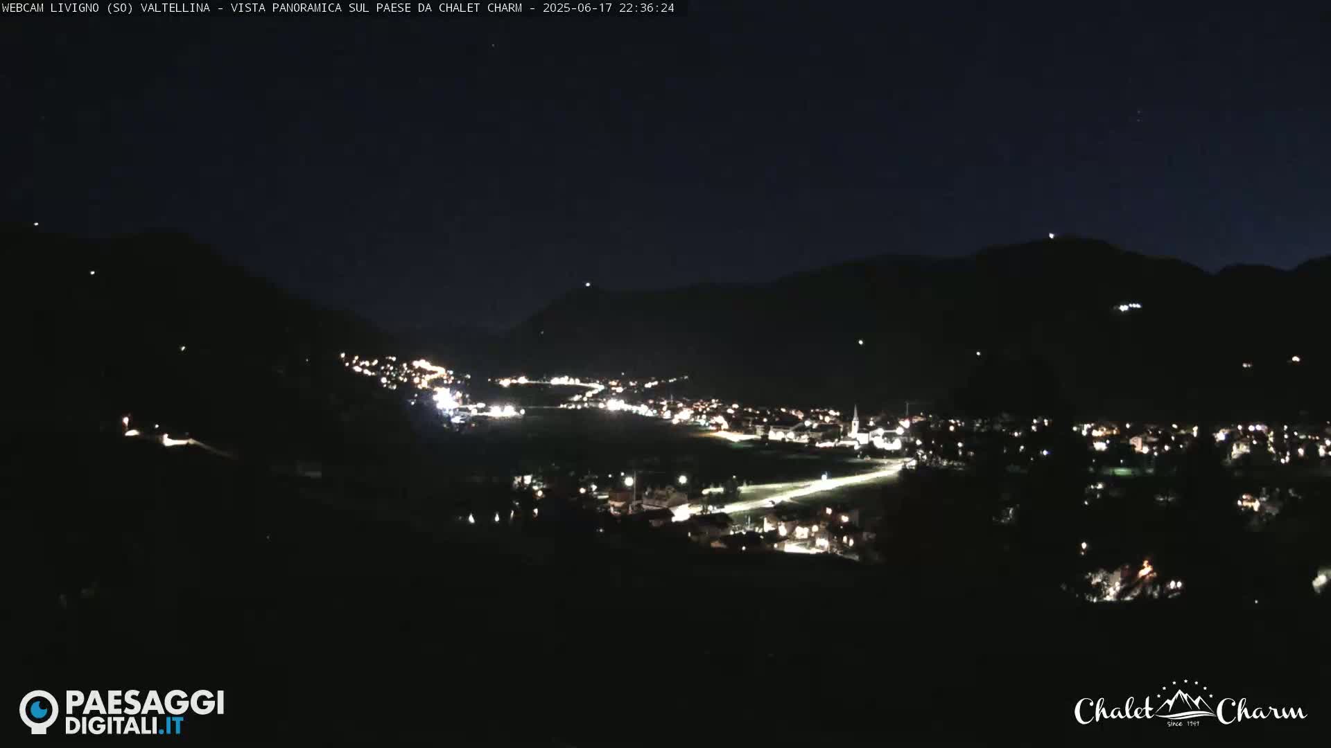 A nighttime panoramic view of a village nestled in a valley between dark mountains under a clear sky.