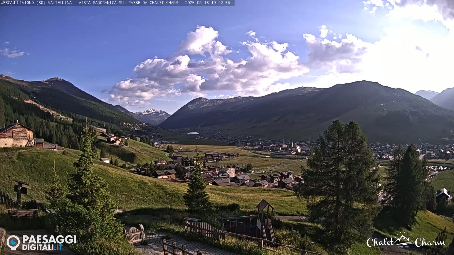 A partly cloudy afternoon scene overlooks a valley town nestled amongst green hills and mountains.