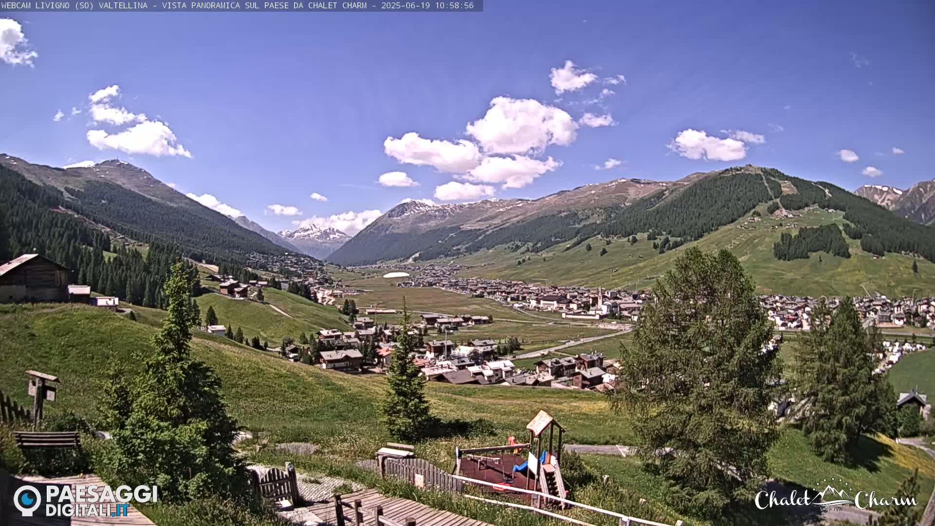 A sunny day reveals a picturesque mountain village nestled in a valley, surrounded by lush green hills and snow-capped mountains under a partly cloudy sky.