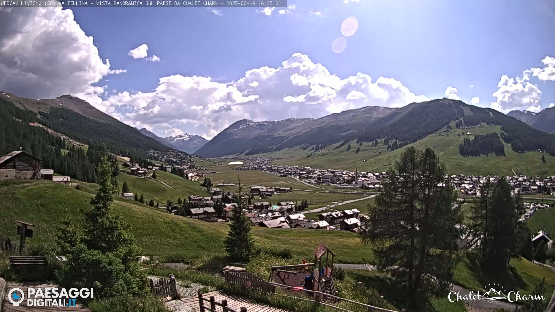 A sunny day with partly cloudy skies overlooks a village nestled in a valley between green hills and mountains.