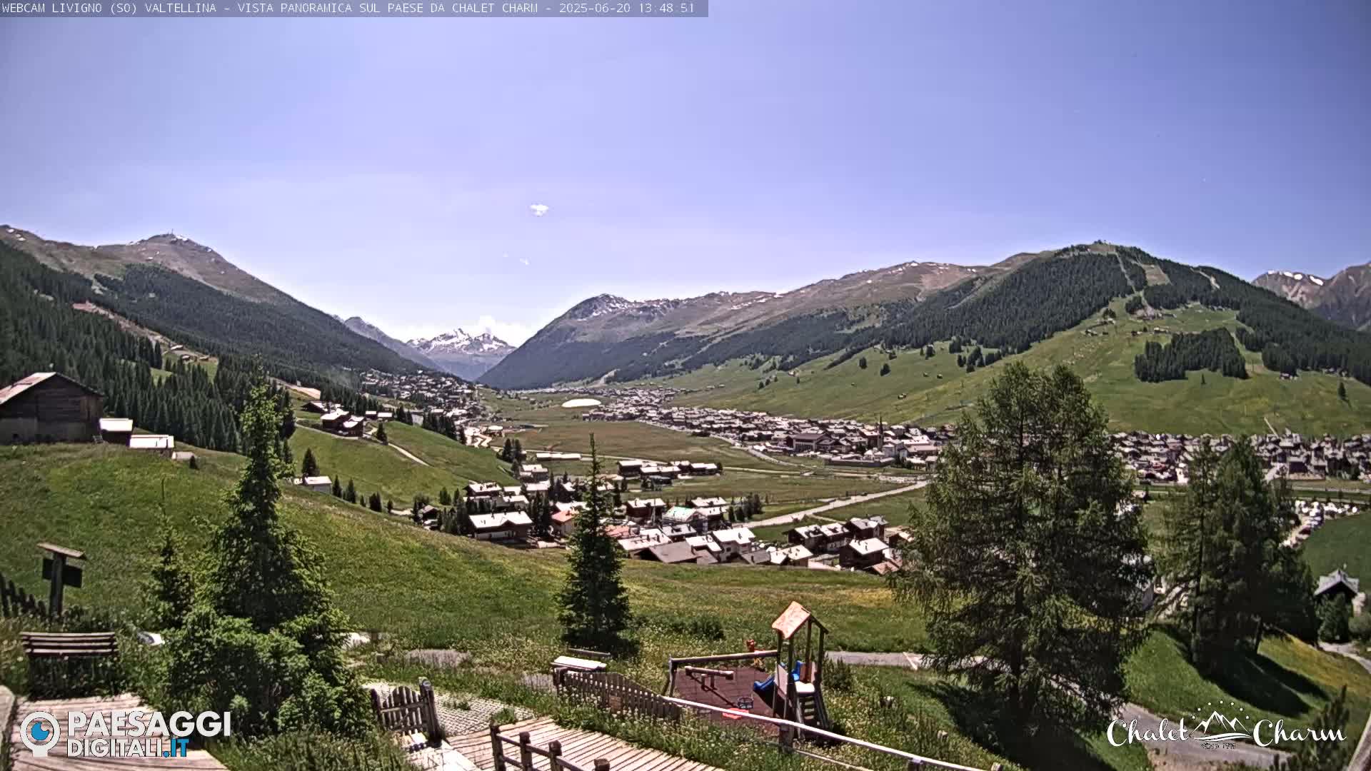 A sunny day reveals a panoramic view of a village nestled in a verdant valley, surrounded by majestic mountains.