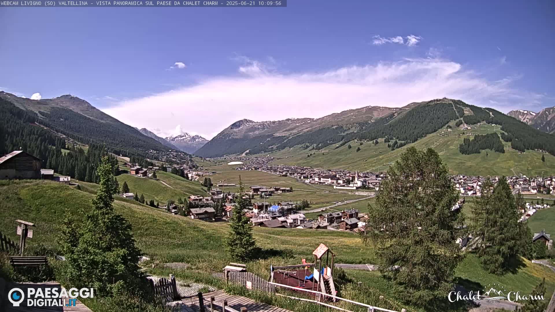 A sunny day reveals a picturesque mountain village nestled in a valley, surrounded by green hills and snow-capped peaks.