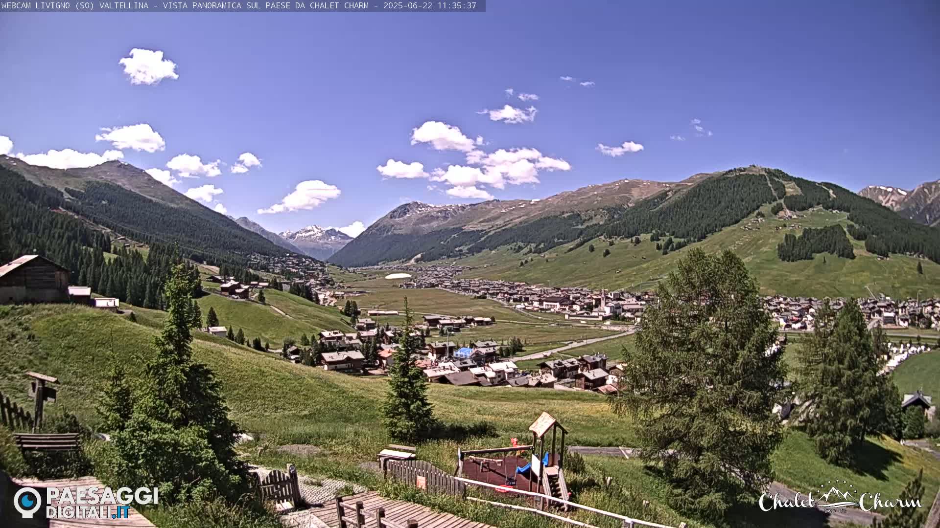A sunny day reveals a sprawling mountain village nestled in a valley, surrounded by verdant hills and snow-capped peaks under a partly cloudy sky.