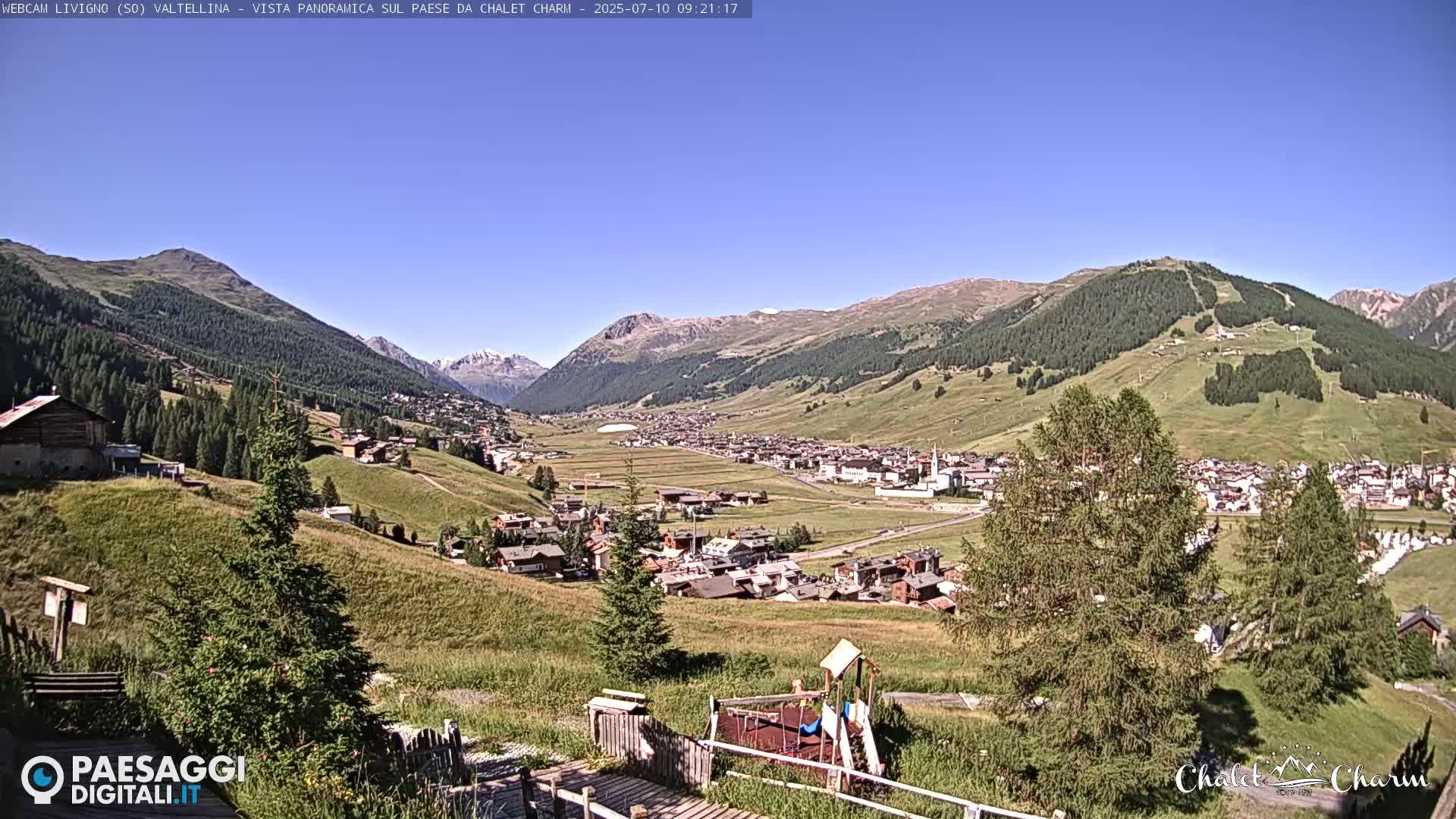 A panoramic view of a valley town nestled among green hills and mountains under a clear blue sky.
