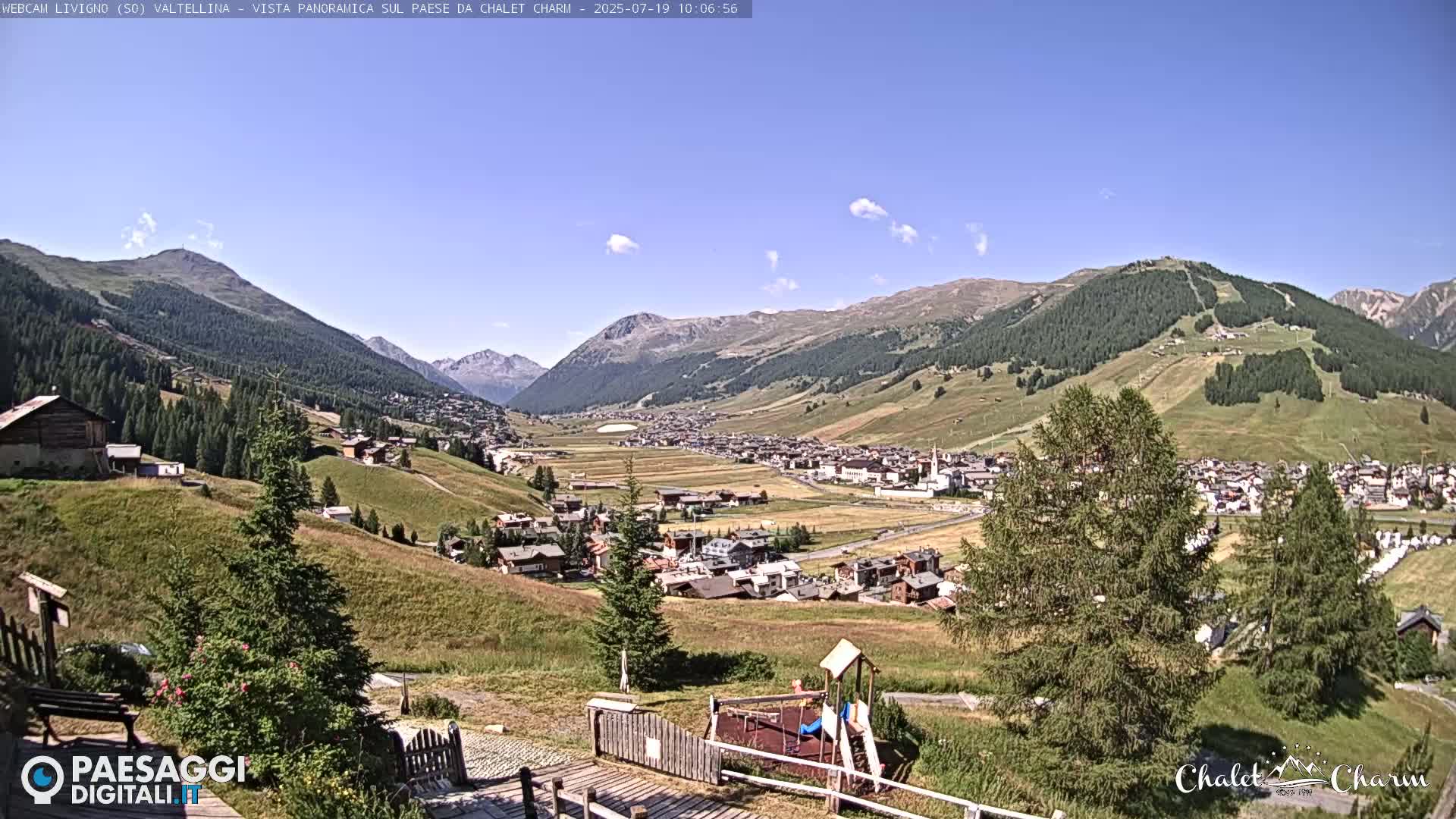 A sunny day reveals a panoramic view of a valley town nestled among rolling green hills and mountains.