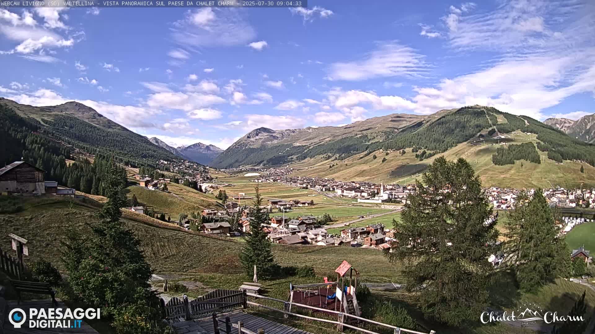 A panoramic view of a valley town nestled among mountains under a mostly sunny sky with scattered clouds.