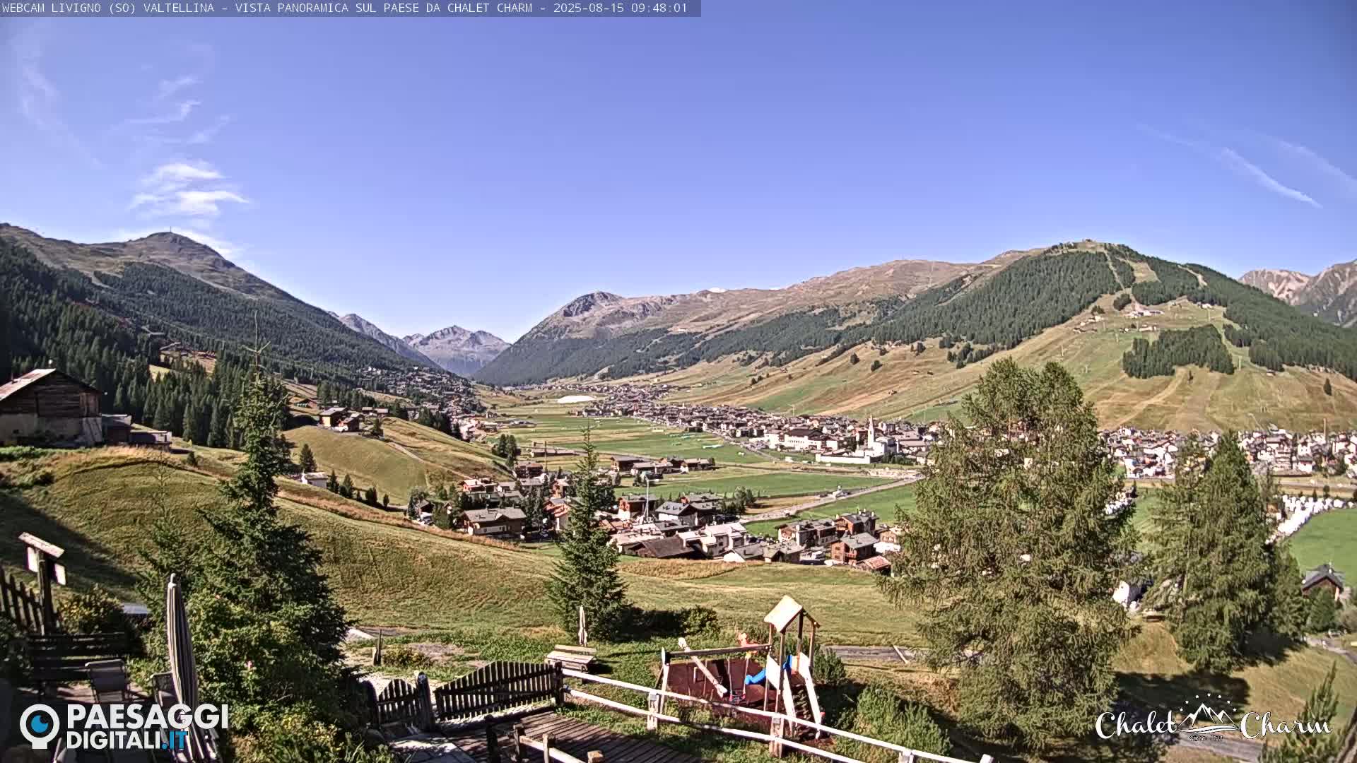 A panoramic view of a valley town nestled between mountains on a clear, sunny day, with a playground visible in the foreground.