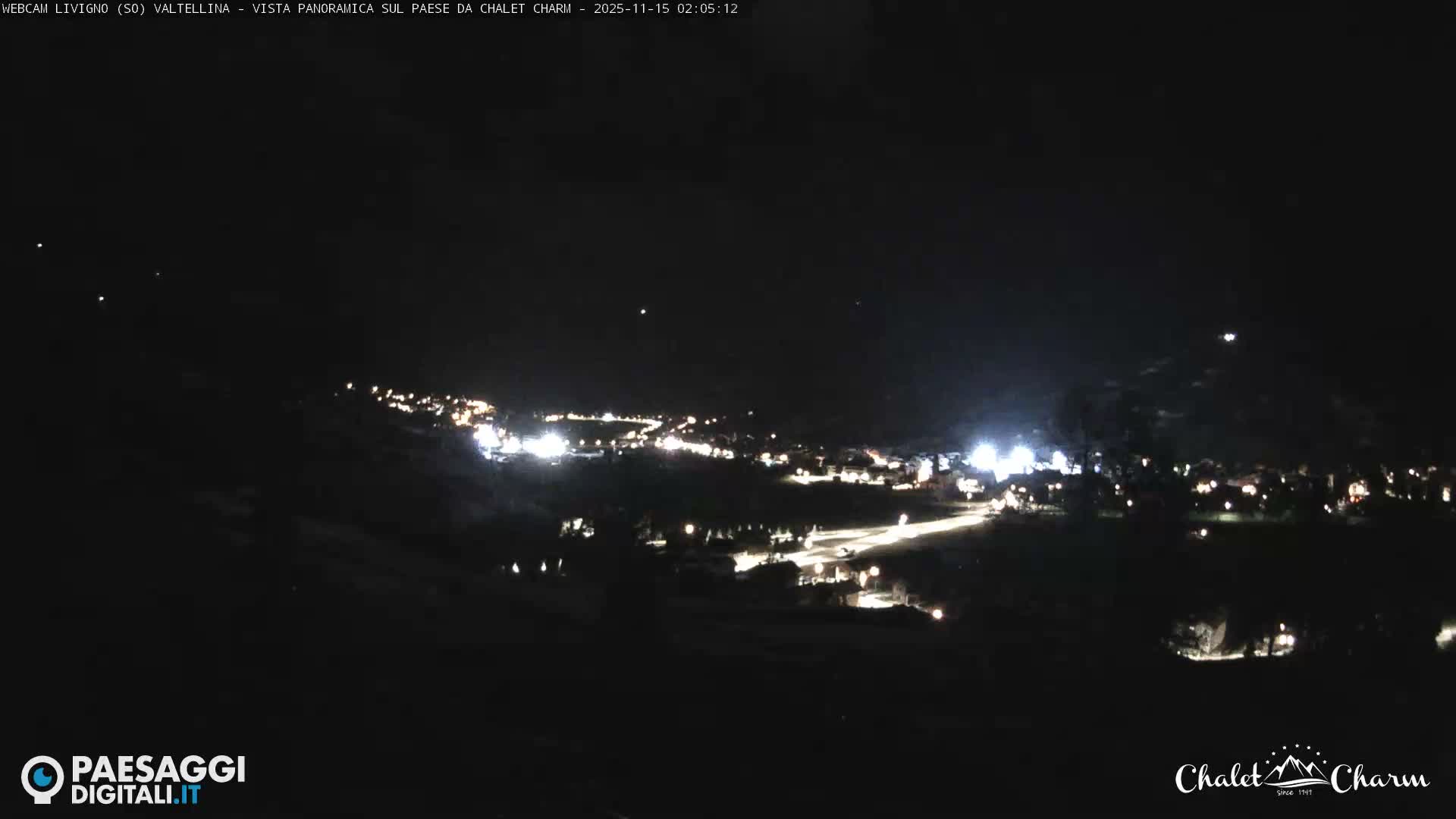 A panoramic nighttime view reveals a sprawling town or village illuminated by numerous artificial lights across a dark landscape under a clear sky.