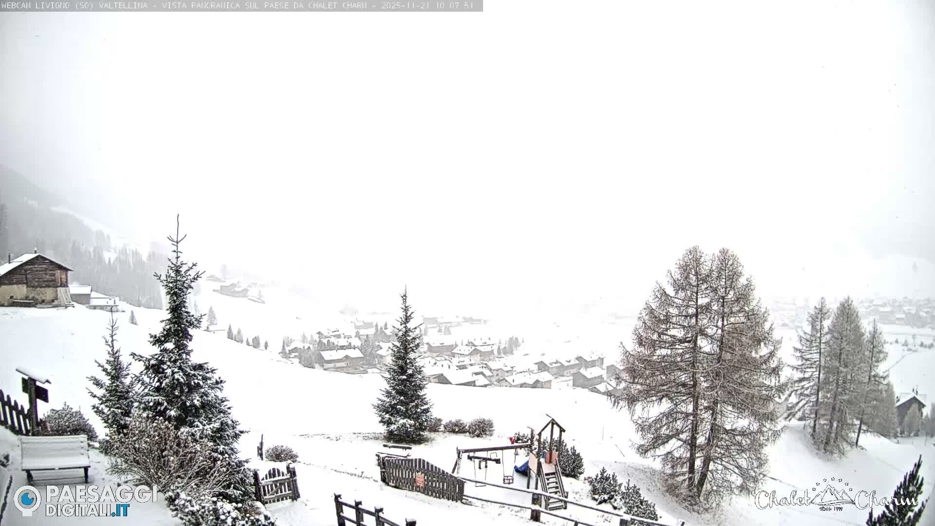 A vast, snow-covered landscape features evergreen trees, a wooden fence, a bench, and a playground in the foreground, leading to a sprawling village nestled in snowy hills under a bright, heavily overcast winter sky.