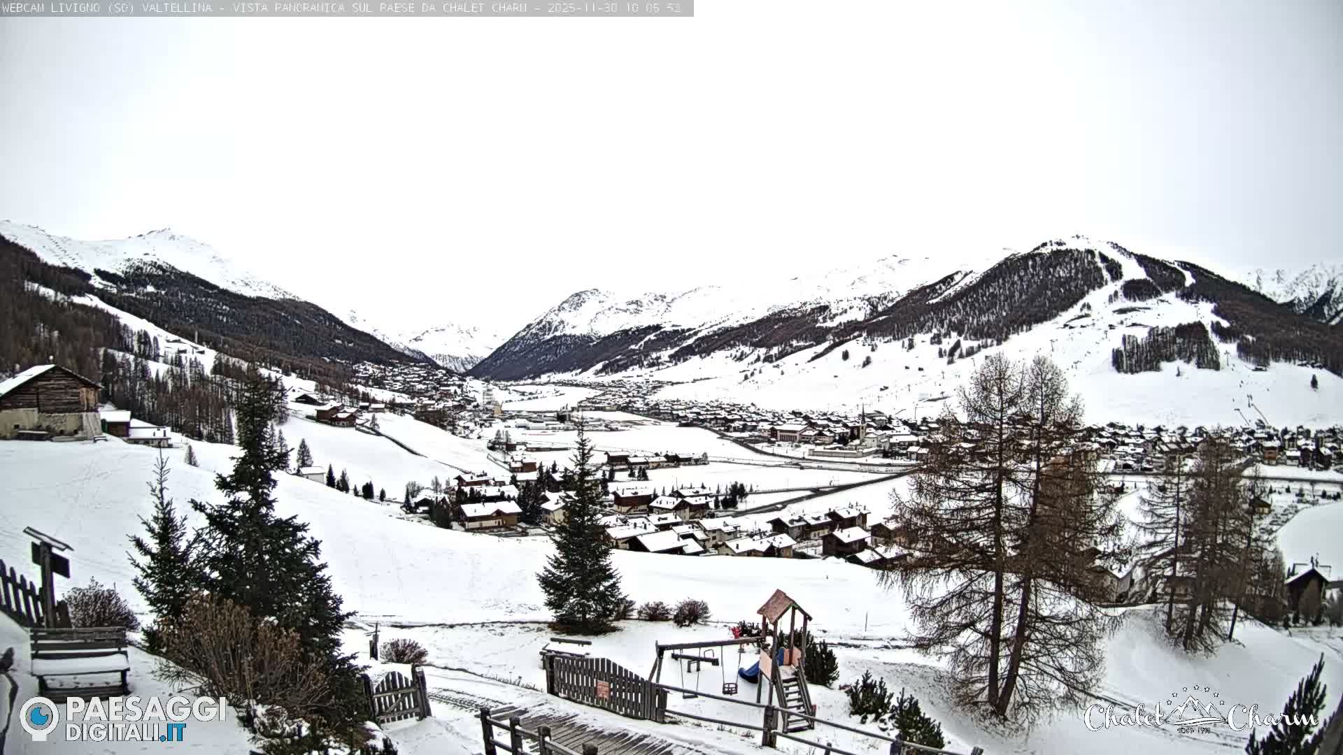 A panoramic view reveals a snow-covered alpine village nestled in a valley surrounded by majestic, snow-capped mountains with forested slopes and visible ski trails, all under an overcast sky, with a children's playground visible in the foreground.