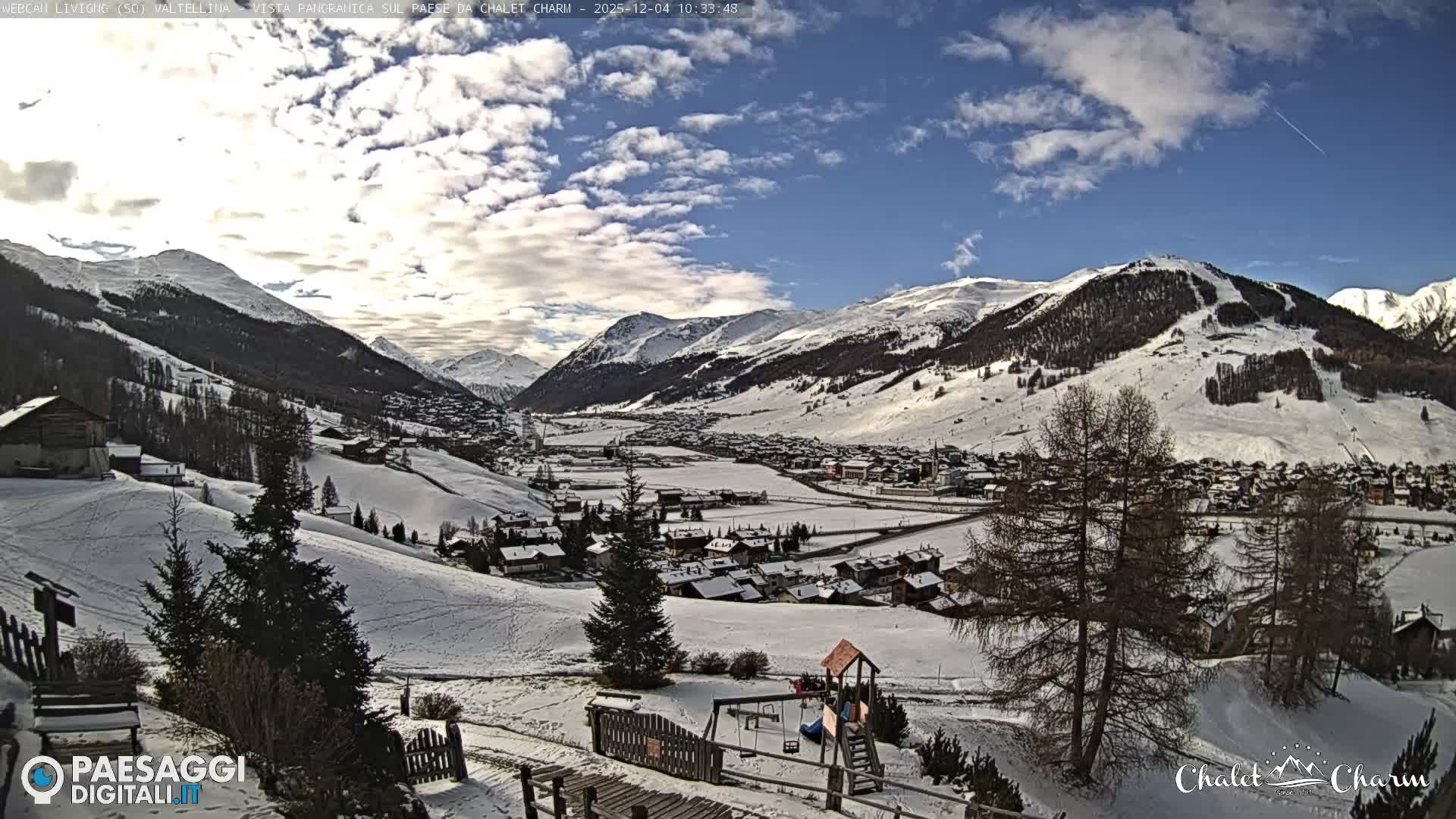 A panoramic view of a snow-covered alpine village nestled in a wide valley, surrounded by majestic mountains featuring visible ski slopes, is seen under a bright blue sky with scattered white clouds on a clear winter day, with a small playground visible in the foreground.