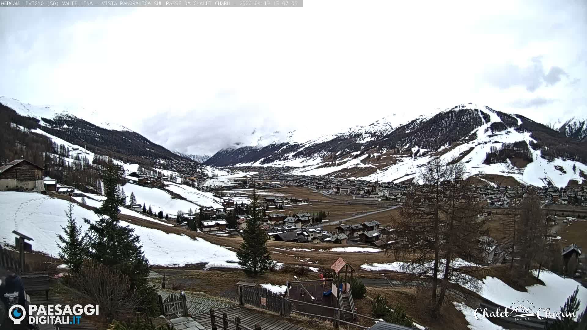A panoramic view of a snow-covered alpine village nestled in a wide valley, surrounded by majestic mountains featuring visible ski slopes, is seen under a bright blue sky with scattered white clouds on a clear winter day, with a small playground visible in the foreground.