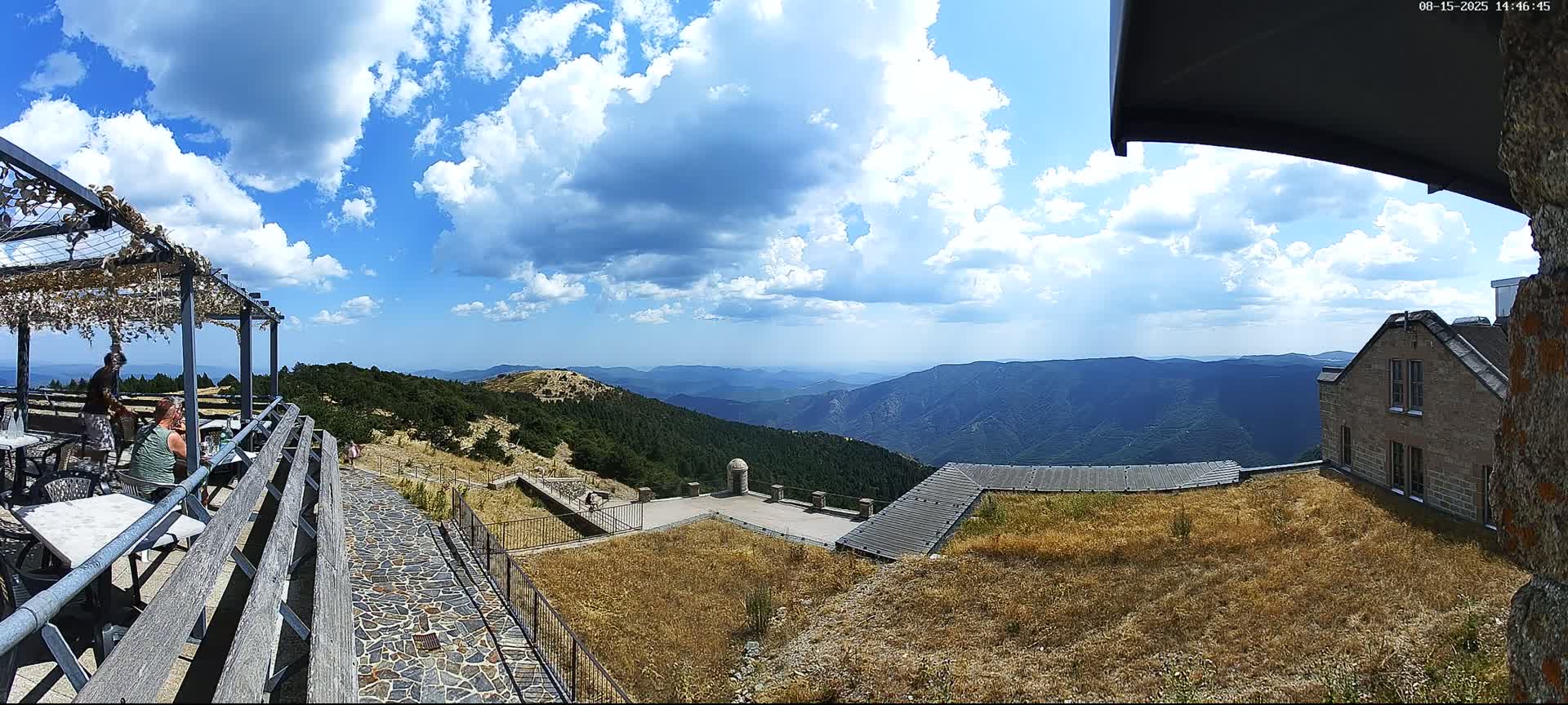 A mountaintop view on a partly cloudy day shows a stone building, an outdoor patio with seating, and expansive mountain ranges in the distance.