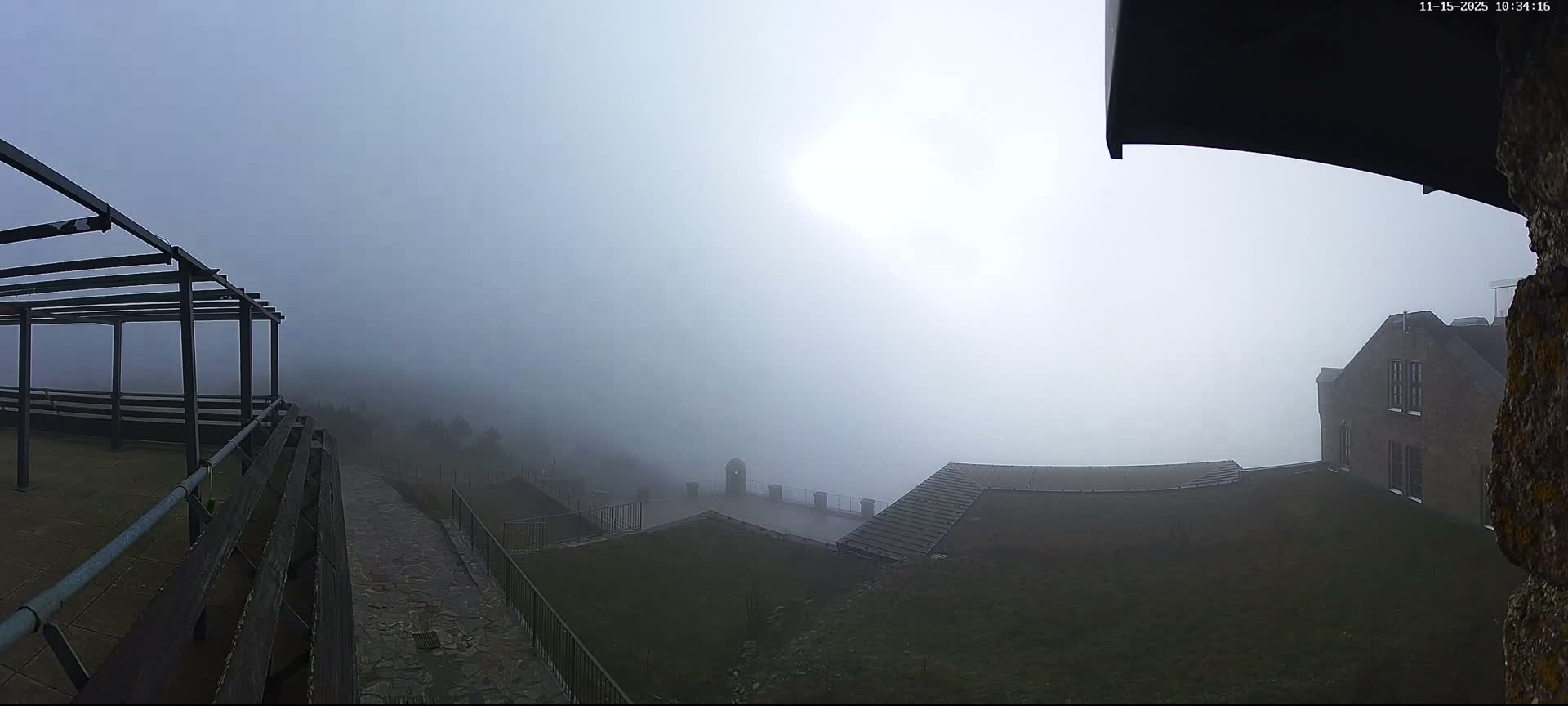 The outdoor scene is heavily obscured by very dense fog, revealing a dark metal pergola and wooden railings on the left, a large brick building on the right, and paved paths with grassy areas in the foreground.