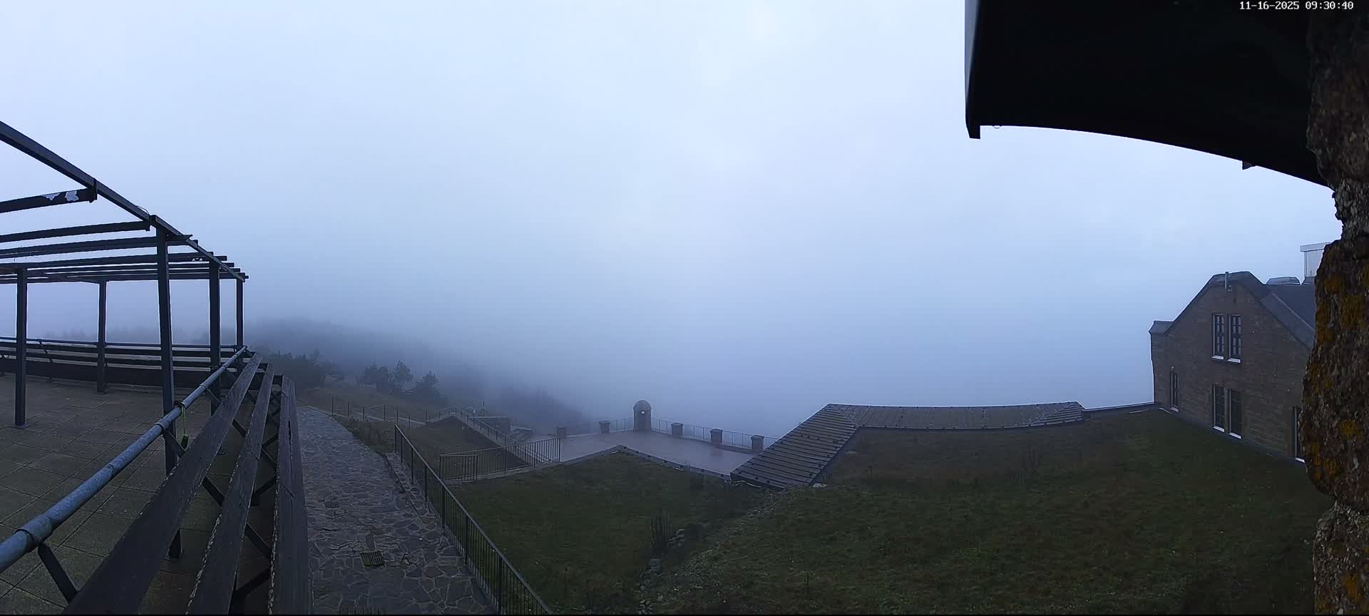 A vast outdoor scene, featuring a metallic pergola, terraced walkways, grassy areas, and a partially visible brick building, is heavily shrouded in dense fog.