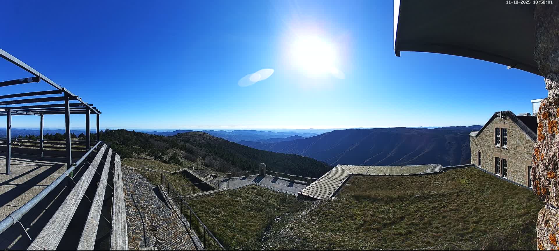 A vast outdoor scene, featuring a metallic pergola, terraced walkways, grassy areas, and a partially visible brick building, is heavily shrouded in dense fog.