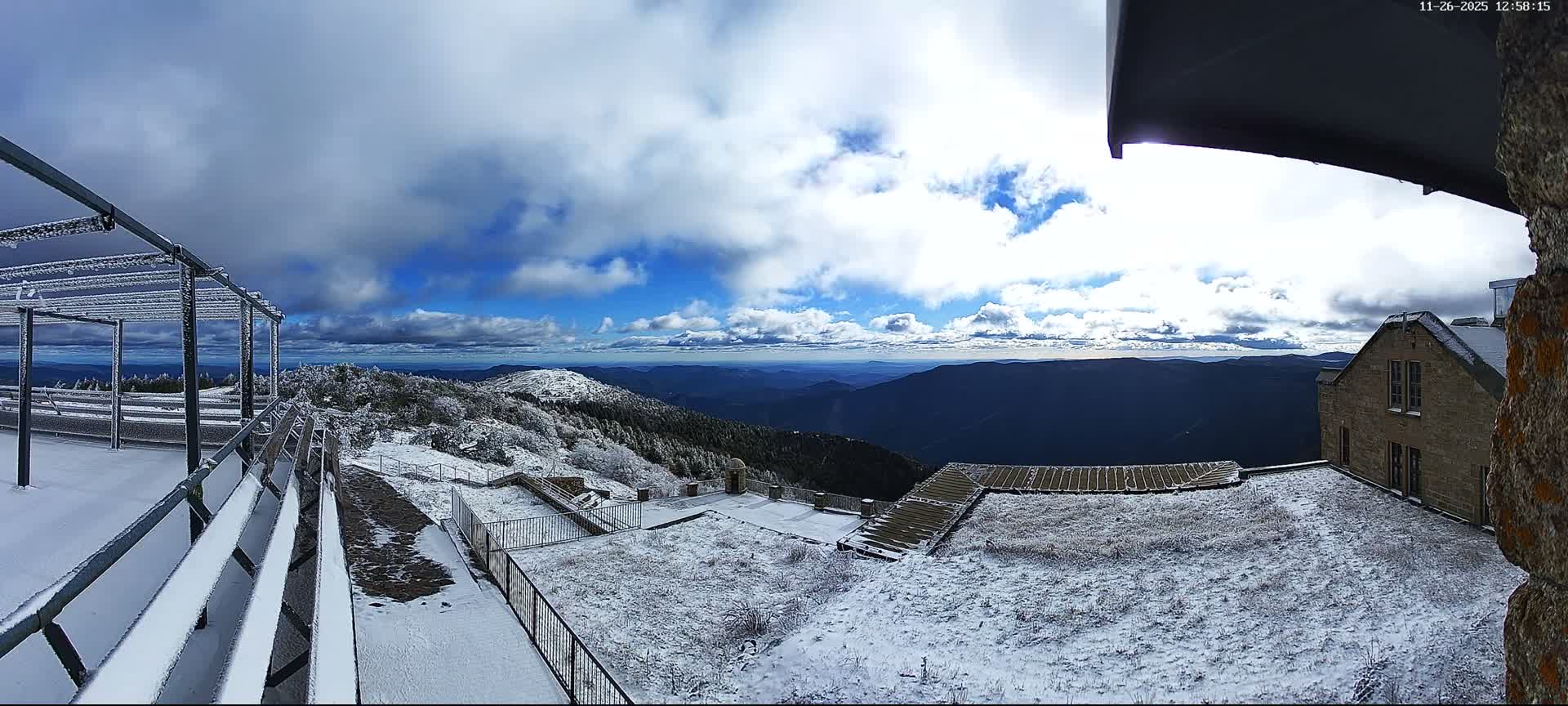 A dense fog envelops an outdoor landscape featuring a dark metal pergola over a wet paved patio on the left, sloping grassy hills disappearing into the haze, and a brick building partially visible on the right, all under heavily overcast and misty conditions.