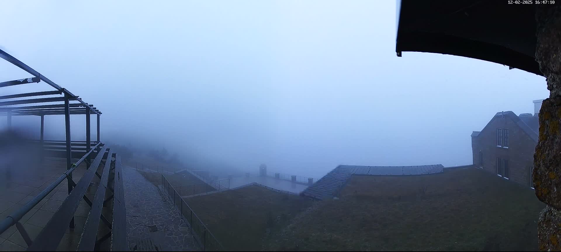 A dense fog shrouds an outdoor area featuring a wet stone-paved path, a dark metal pergola and railings, a grassy hillside, and the top of a multi-story building barely visible through the heavy mist.