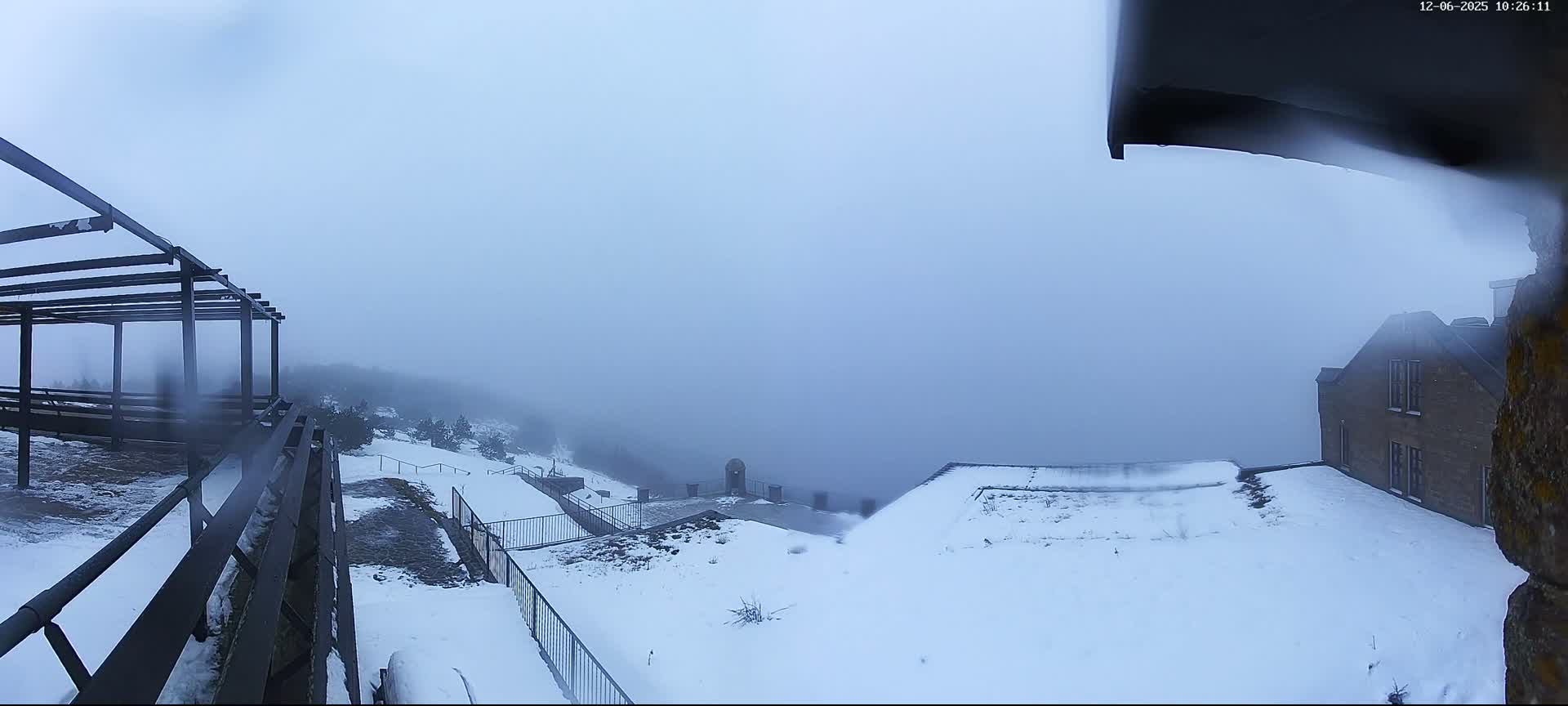The image captures a snow-covered mountainous landscape blanketed in dense fog, featuring a stone building and dark metal structures under a cold, overcast sky.